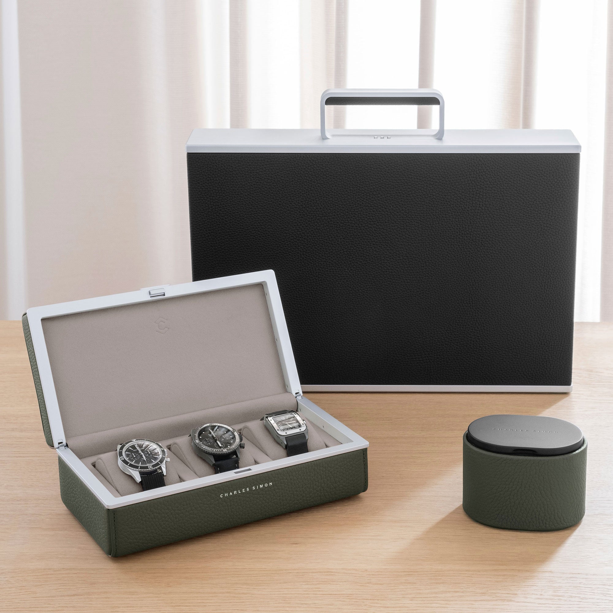 Charles Simon watch accessories collection in khaki leather. An open Eaton 3 watch case with 3 luxury watches inside is open on the left, a closed Theo is placed on the right and a Mackenzie watch briefcase is placed in the center in the back. 