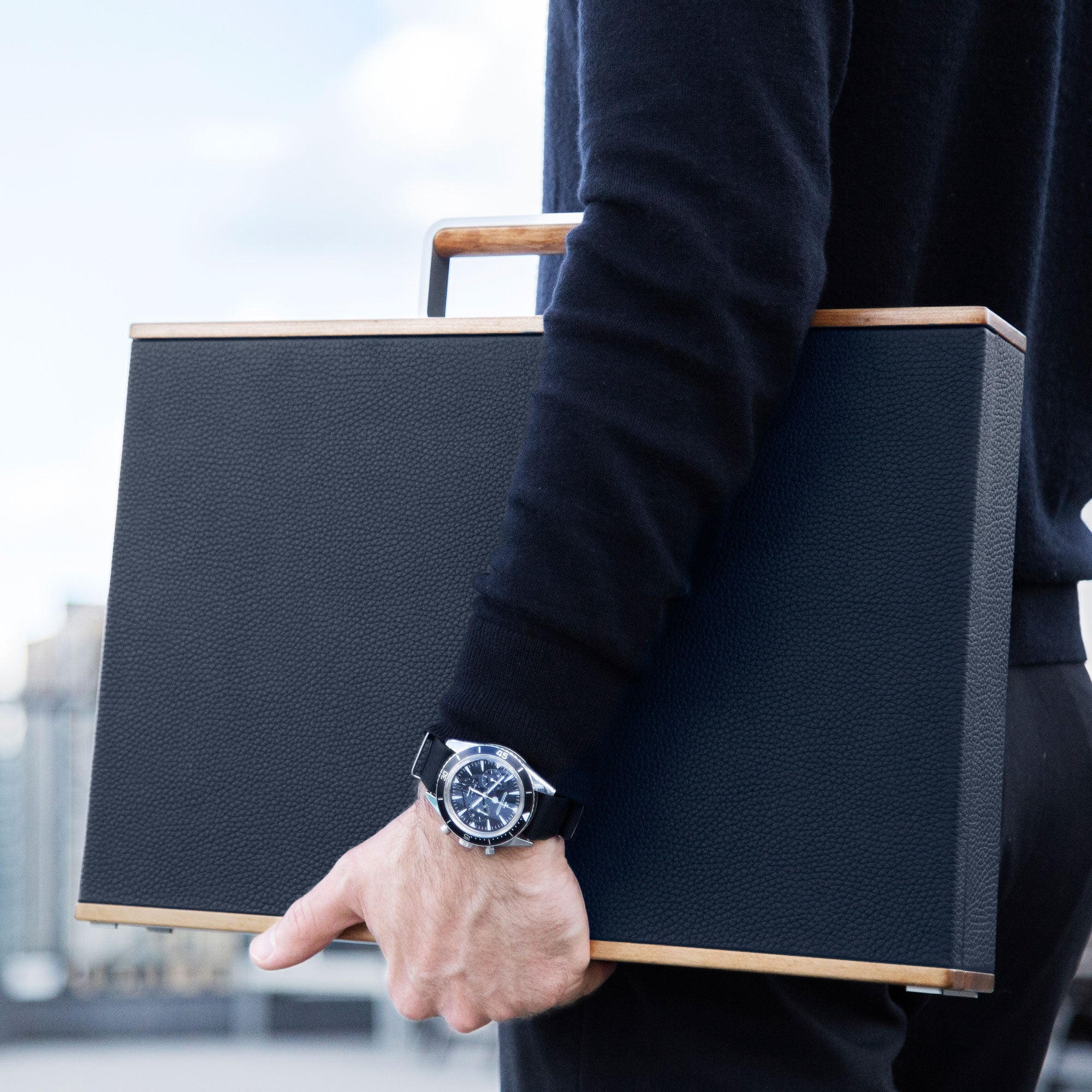 Lifestyle photo of man wearing luxury watch holding Mackenzie Original watch briefcase handmade from recycled wood, young bull leather and carbon fiber and anodized aluminum casing. 