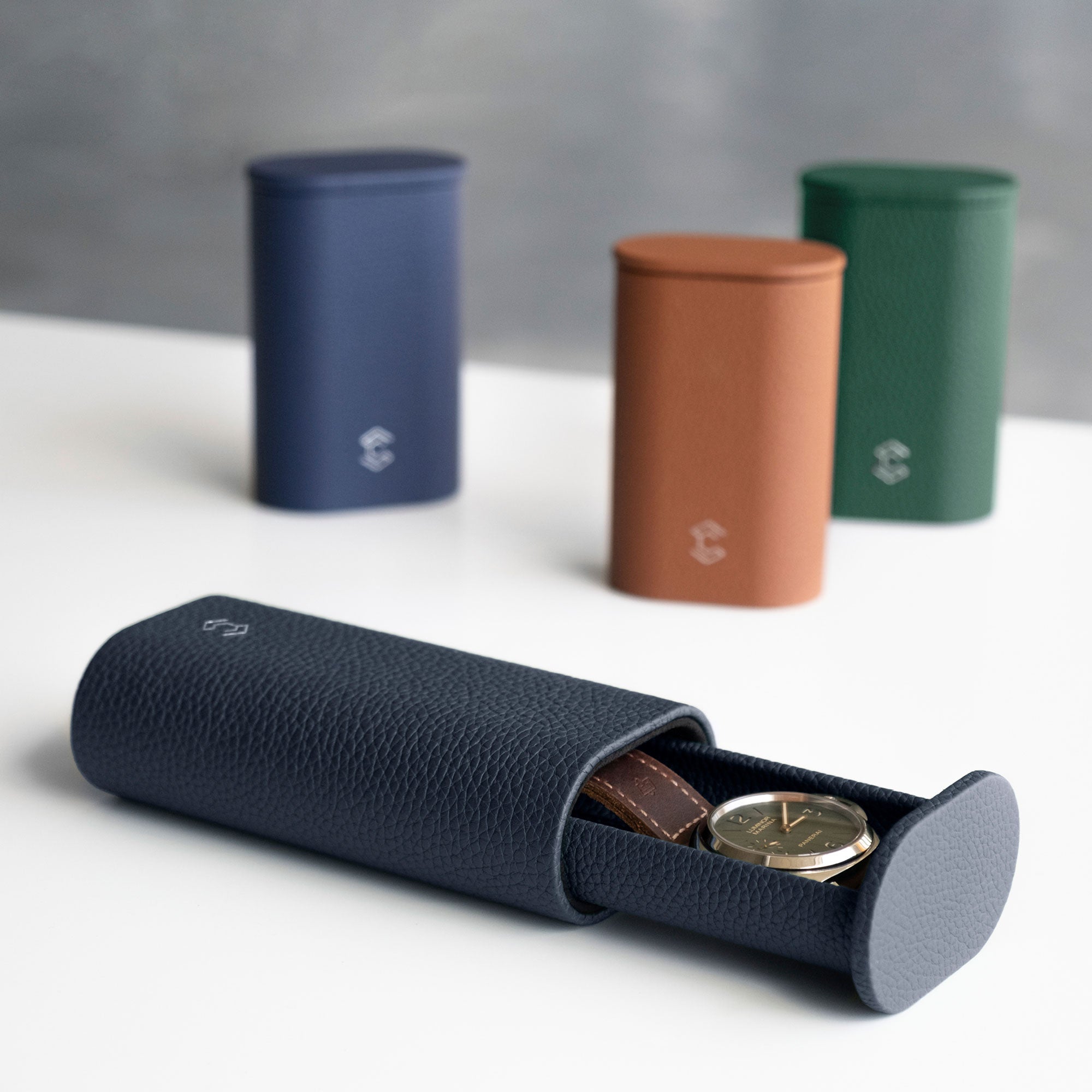 Lifestyle product photo of open Oscar 1 Tall Travel Watch roll in marine leather holding a mens watch with a leather watch strap. Closed Oscar Watch rolls in different shades of French leather are placed standing upright in the background.