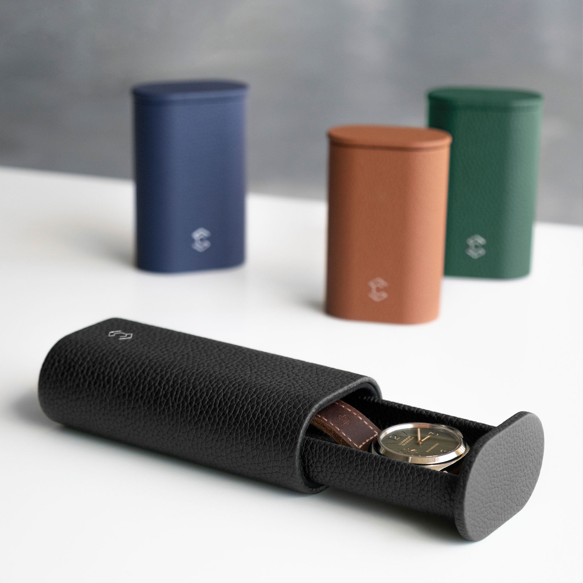 Photo of open Oscar 1 Tall Watch roll in black leather holding a luxury watch with a leather watch strap. Closed Oscar Watch rolls in different shades of French leather are placed standing upright in the background.