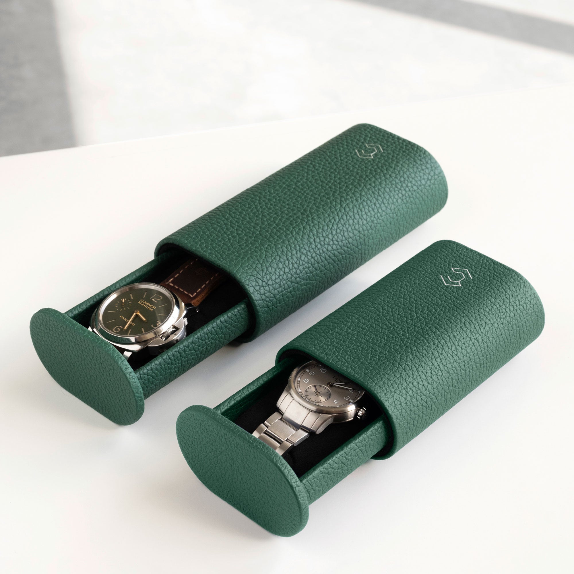 Lifestyle image of Oscar Tall leather watch roll in emerald leather on the left, holding a watch with a leather strap and a half opened emerald Oscar Watch roll for watches with metal bracelets on the right