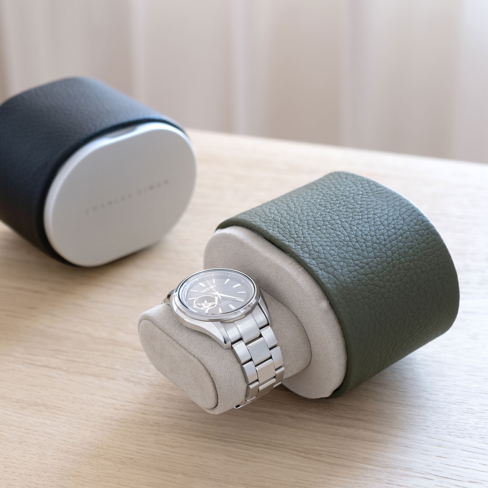 Lifestyle shot of open khaki Theo leather watch roll holding one watch on sea sand Alcantara watch cushion. Closed marine Theo luxury watch roll is placed in the background.