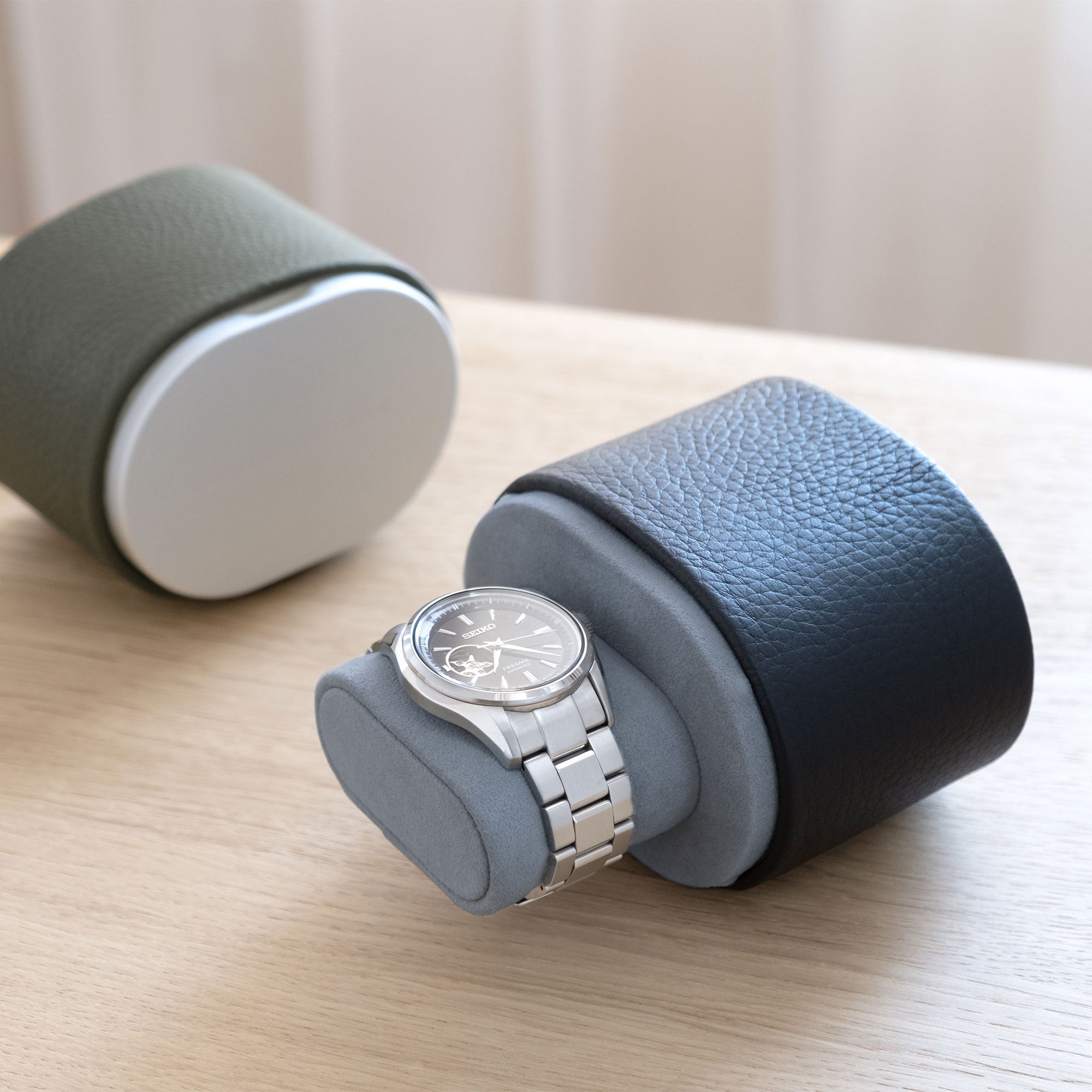 Lifestyle shot of open marine leather Theo watch roll holding one watch on sea sand Alcantara watch cushion. Closed khaki Theo luxury watch roll is placed in the background.