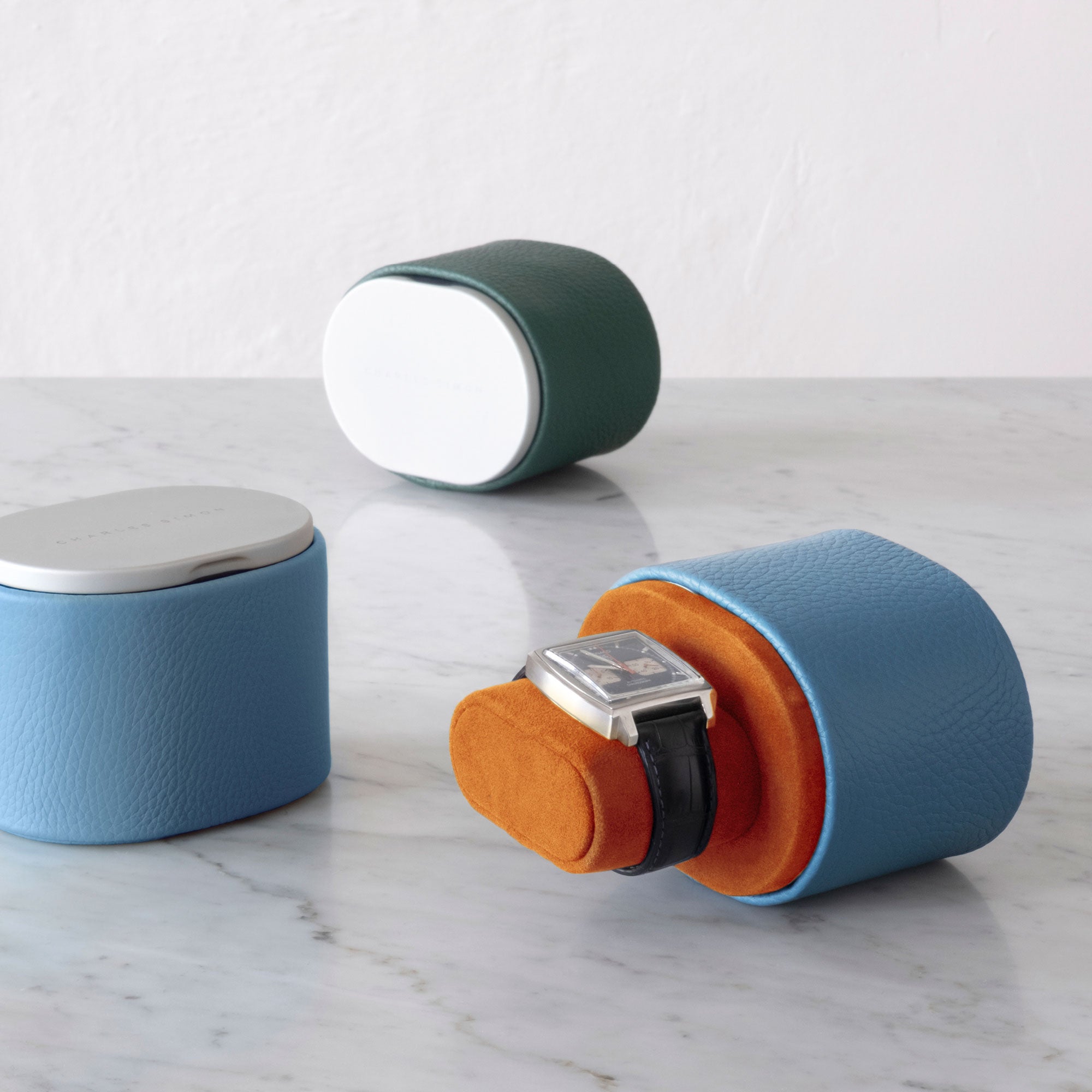 Lifestyle photo of 2 Theo watch rolls. Watch roll in the foreground is made from sky blue leather and open, placed on marble table with luxury watch on Papaya Alcantara cushion. Theo watch roll in the back is closed and made from emerald leather.