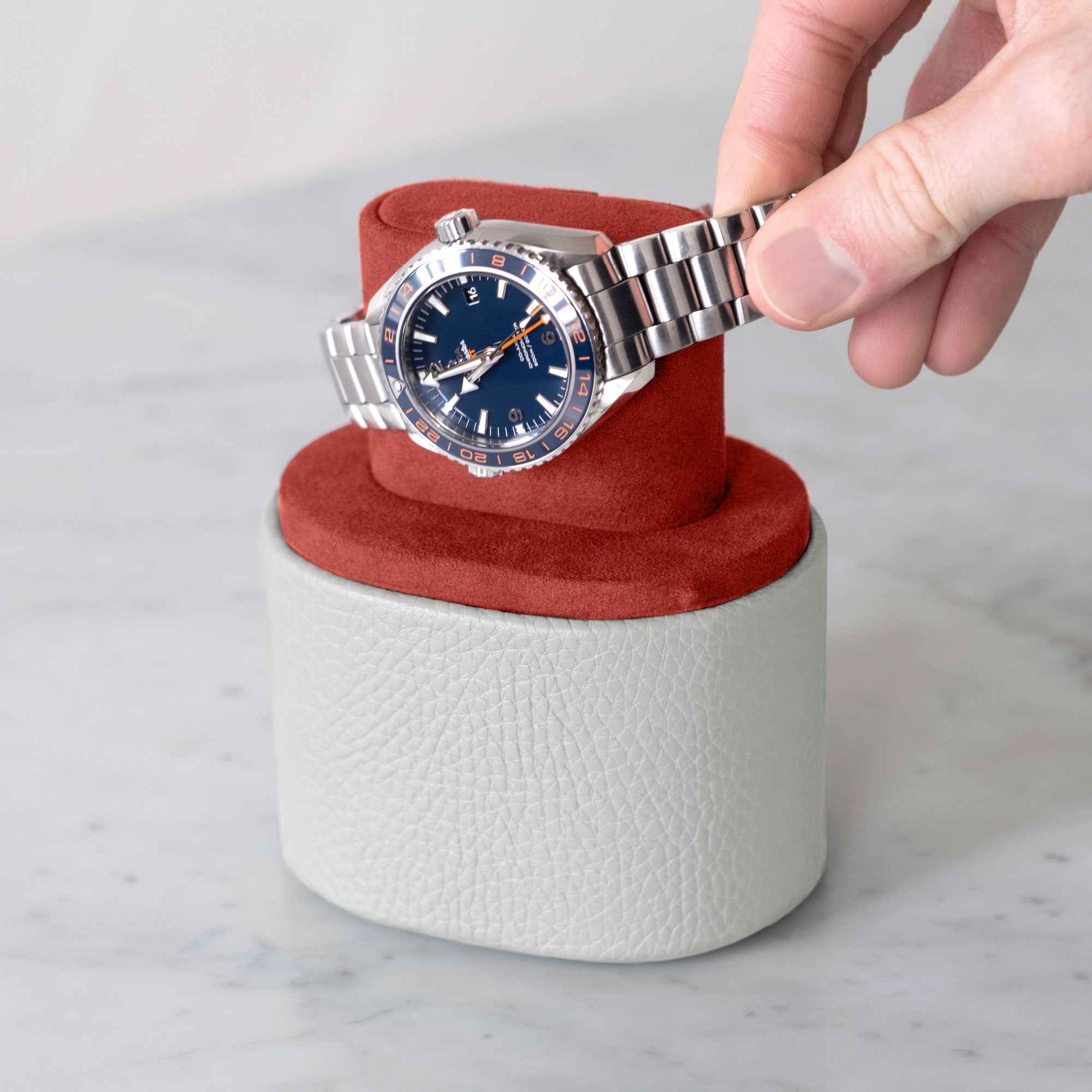 Lifestyle shot of handmade Theo watch roll in white leather and Vermilion interior holding Omega Planet Ocean luxury watch