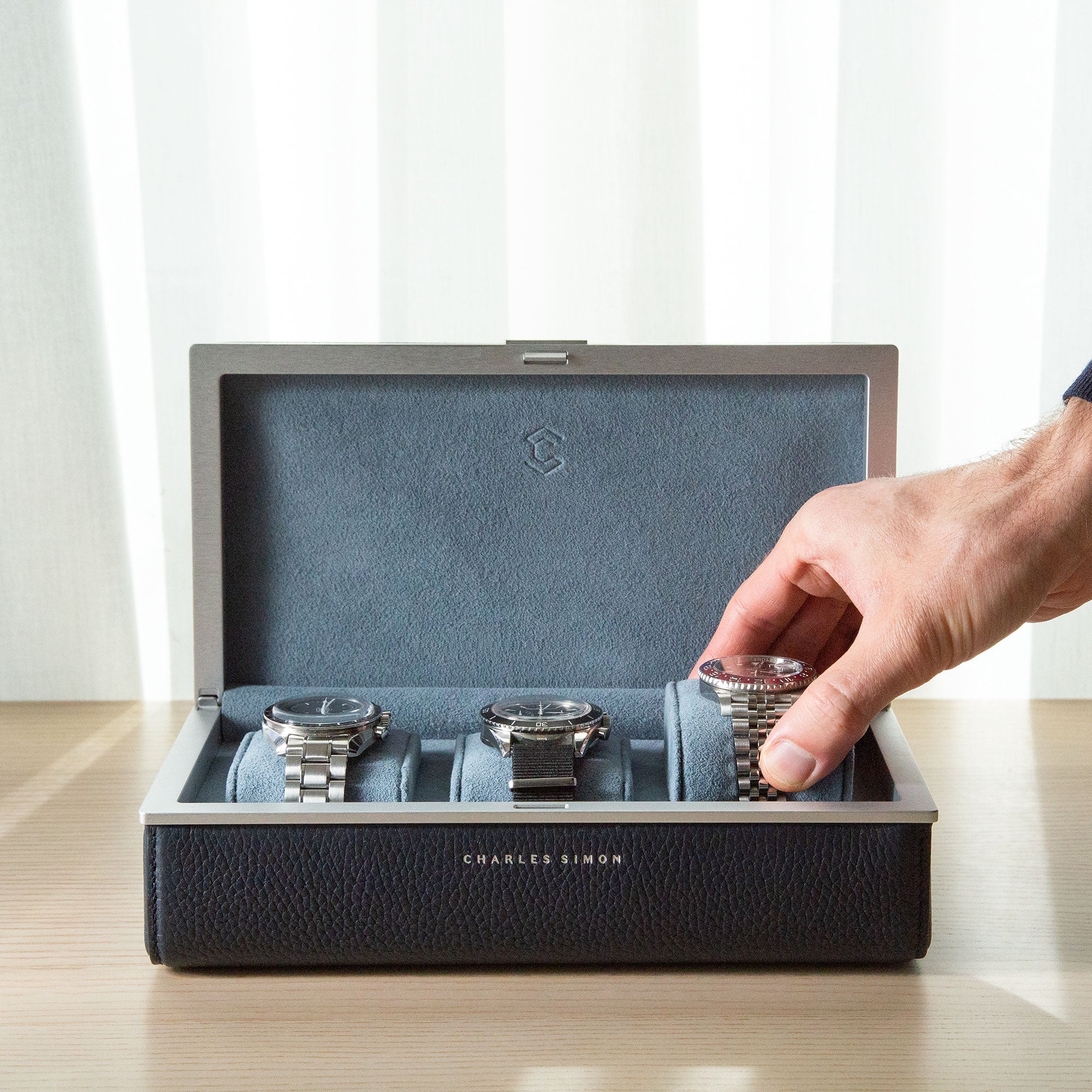 Lifestyle shot of man taking luxury watch placed on blue removable Alcantara cushion from a Charles Simon Eaton 3 watch case in marine that is holding three men's watches