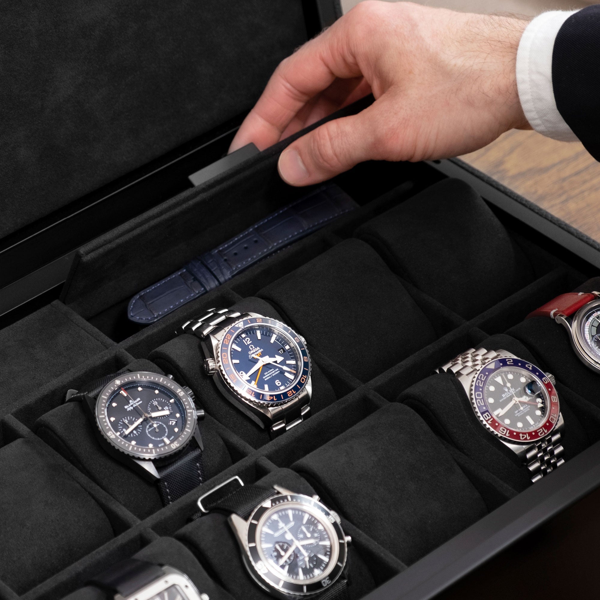 Closeup photo of hand opening additional compartment of the Spence 12 Watch box in all black. The storage compartment is holding an extra leather watch strap. Luxury watches from watch brands including Omega, Jaeger LeCoultre, Rolex and others are displayed on removable Alcantara cushions.