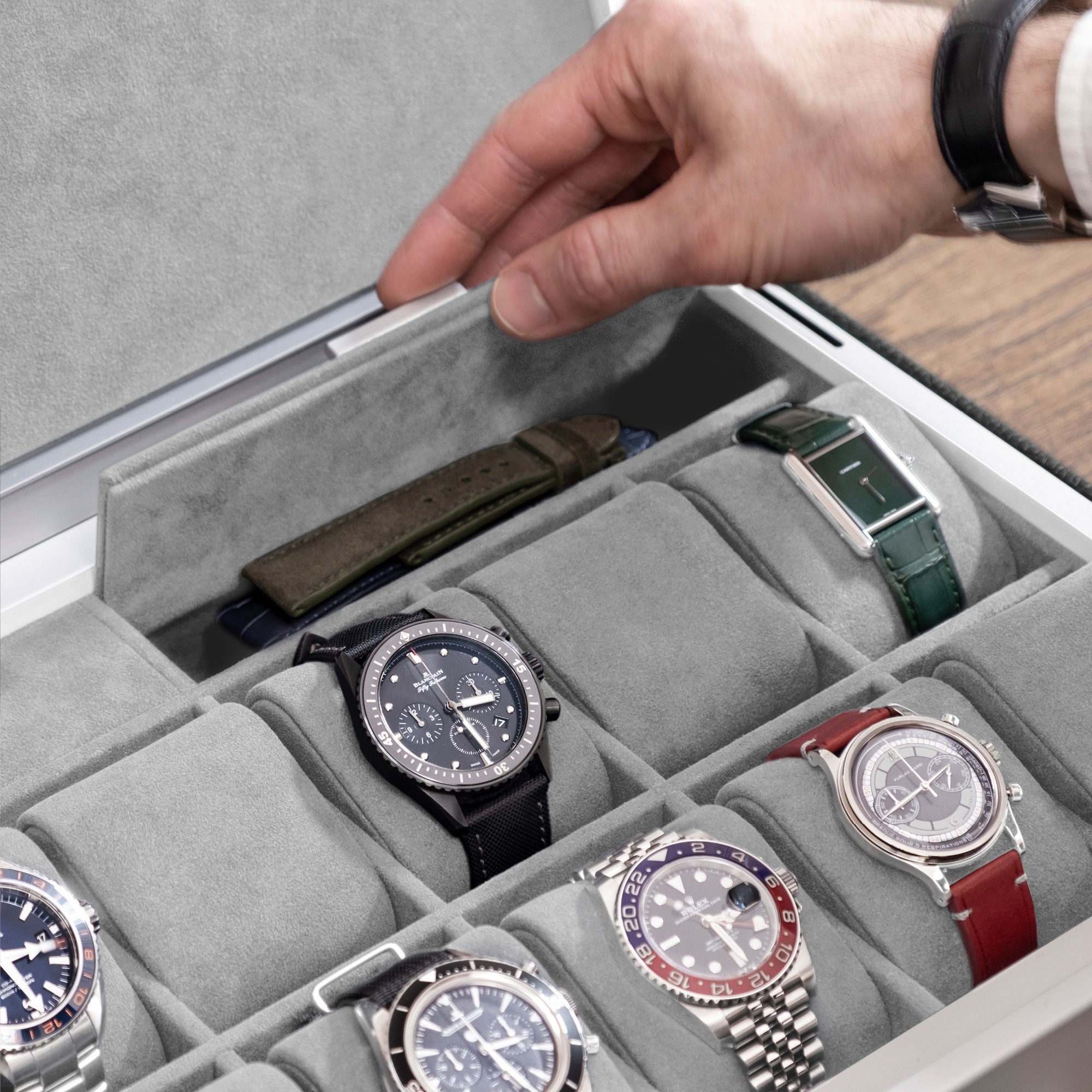 Closeup photo of hand opening additional compartment of the Spence 12 Watch box with fog grey Alcantara interior lining. The storage compartment is holding an extra leather watch strap. Luxury watches from watch brands including Omega, Cartier, Rolex and others are displayed on removable watch cushions.