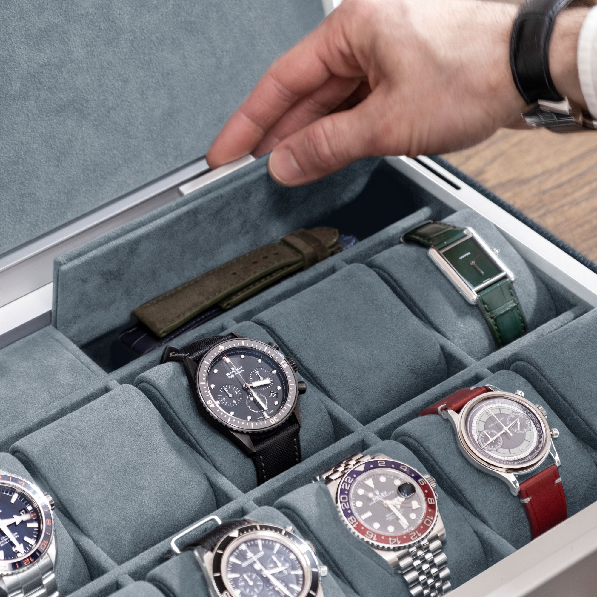 Closeup photo of hand opening additional compartment of the Spence 12 Watch box in marine leather and smoky blue interior. The storage compartment is holding an extra leather watch strap. Luxury watches from watch brands including Omega, Jaeger LeCoultre, Rolex and others are displayed on removable Alcantara cushions.