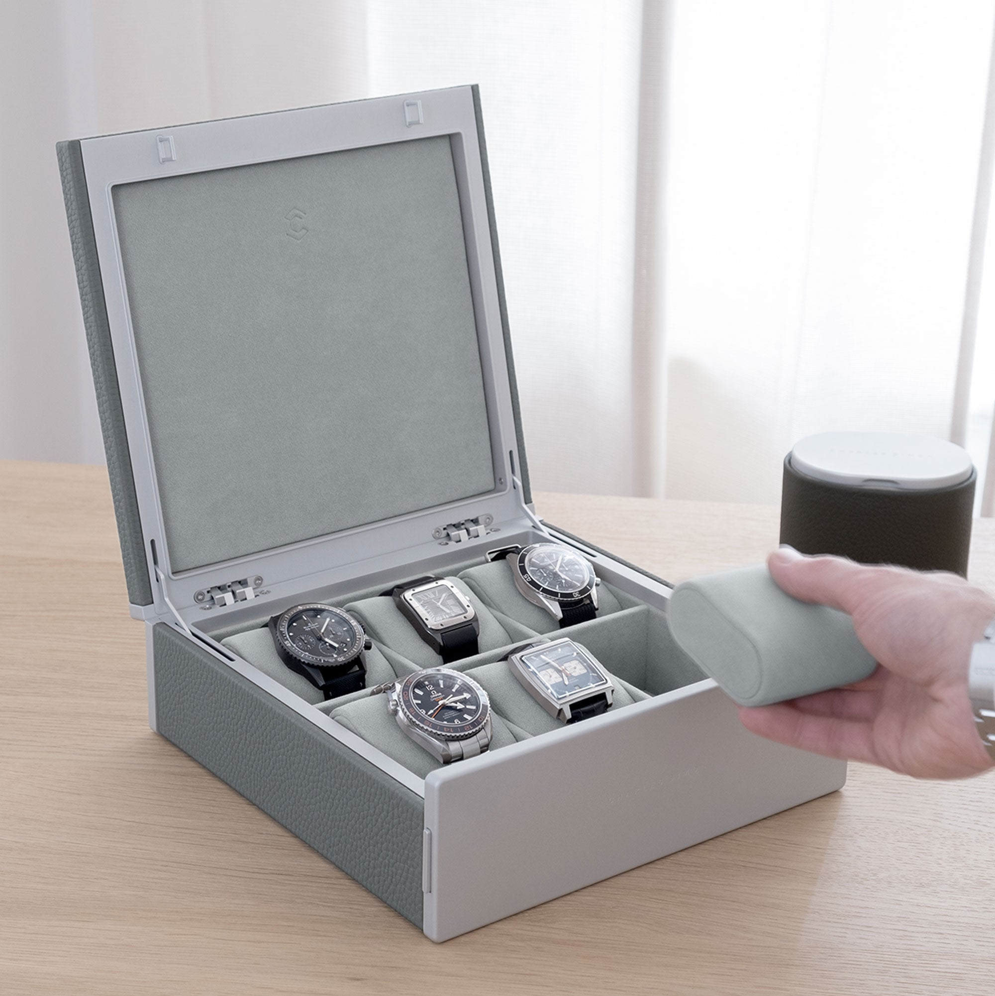 Lifestyle shot of Spence watch box in cloud grey leather and grey carbon fiber and anodized aluminum containing 5 luxury watches including Cartier, Rolex, Jaeger Lecoultre, Seiko and Omega on fog grey removable Alcantara watch cushions. Gentleman is grabbing luxury watch from Theo watch roll in khaki in the background