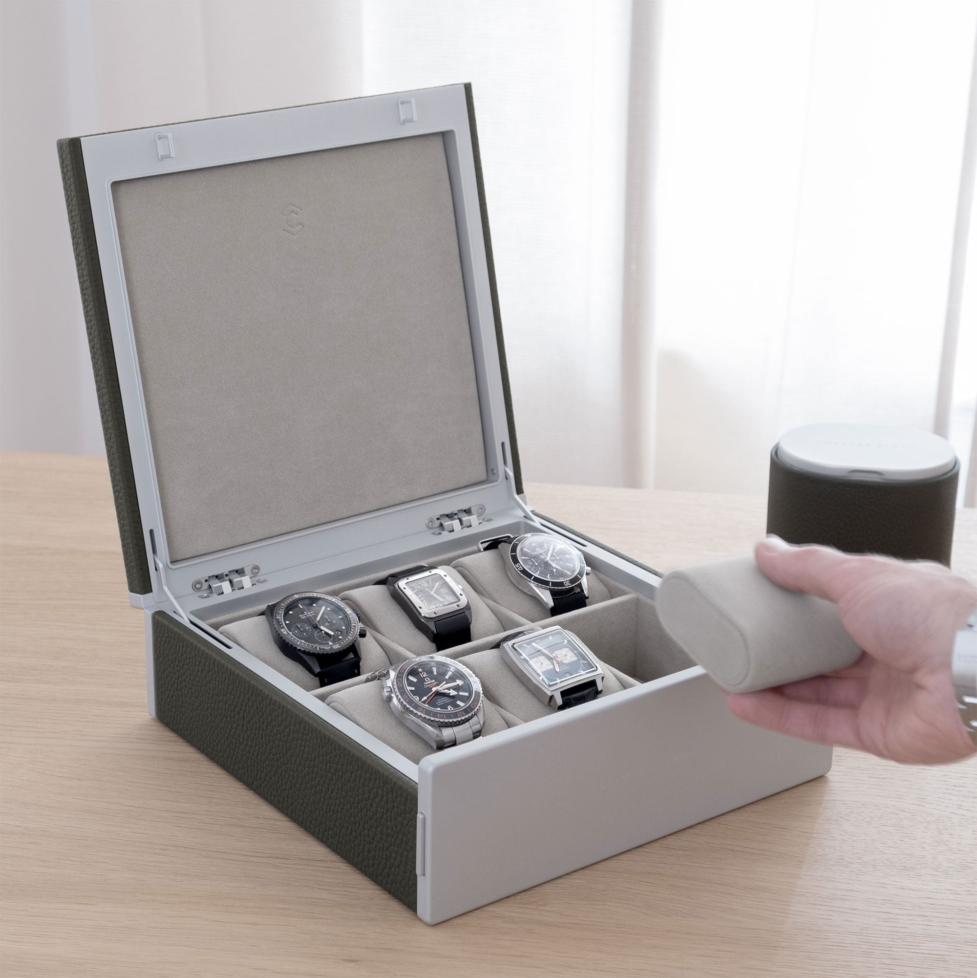 Luxury lifestyle shot of Spence watch box in graphite leather for 6 watches filled with men's luxury watches including Rolex, IWC, Jaeger Lecoultre, Cartier and Omega. Gentleman is taking removable Alcantara cushion from the Spence watch box.