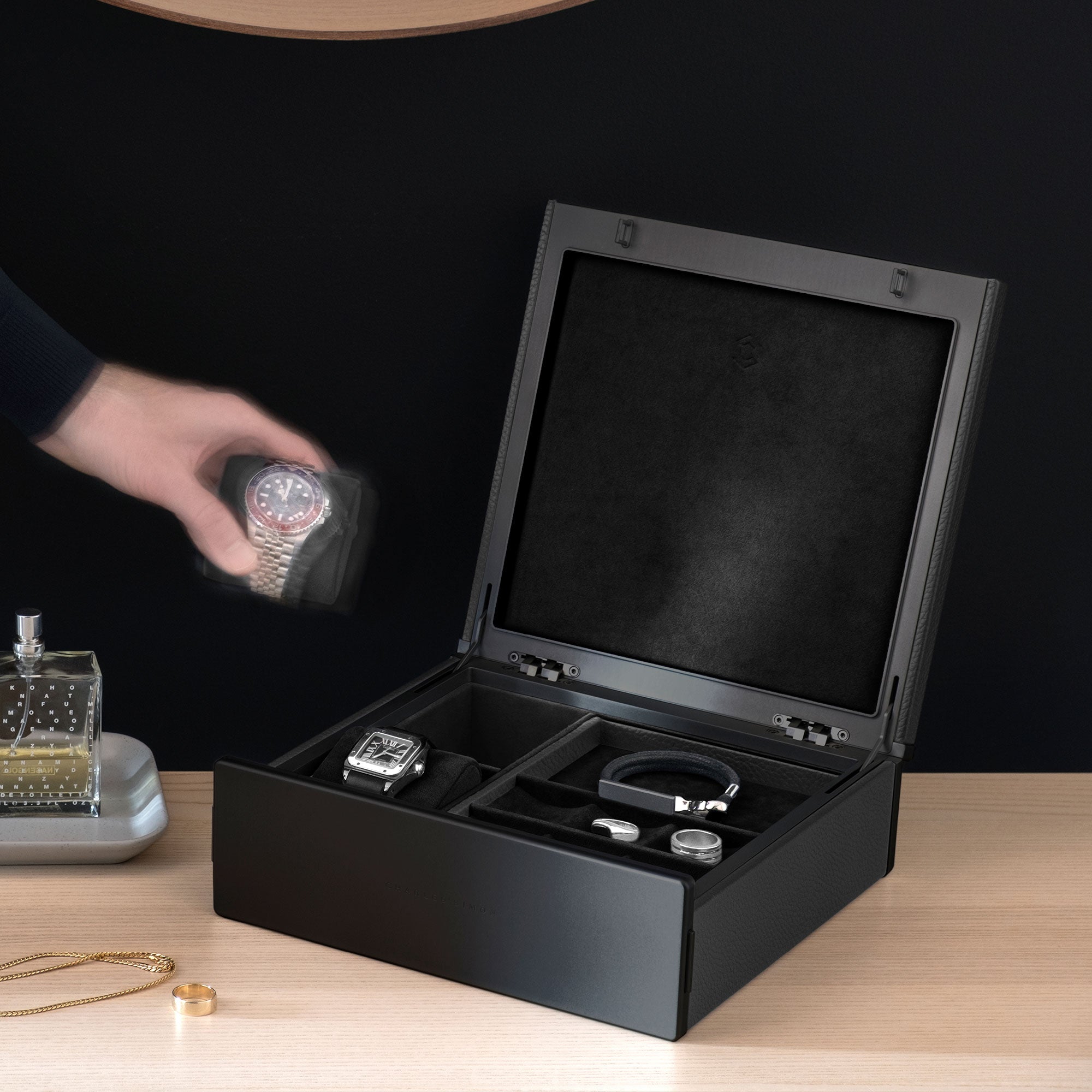 Lifestyle photo of all black Taylor 2 Watch and Jewelry box. Man is taking Rolex watch placed on watch cushion from the watch and jewelry box. The Taylor 2 Watch and Jewelry box holds a black leather bracelet, silver rings and a Cartier watch.