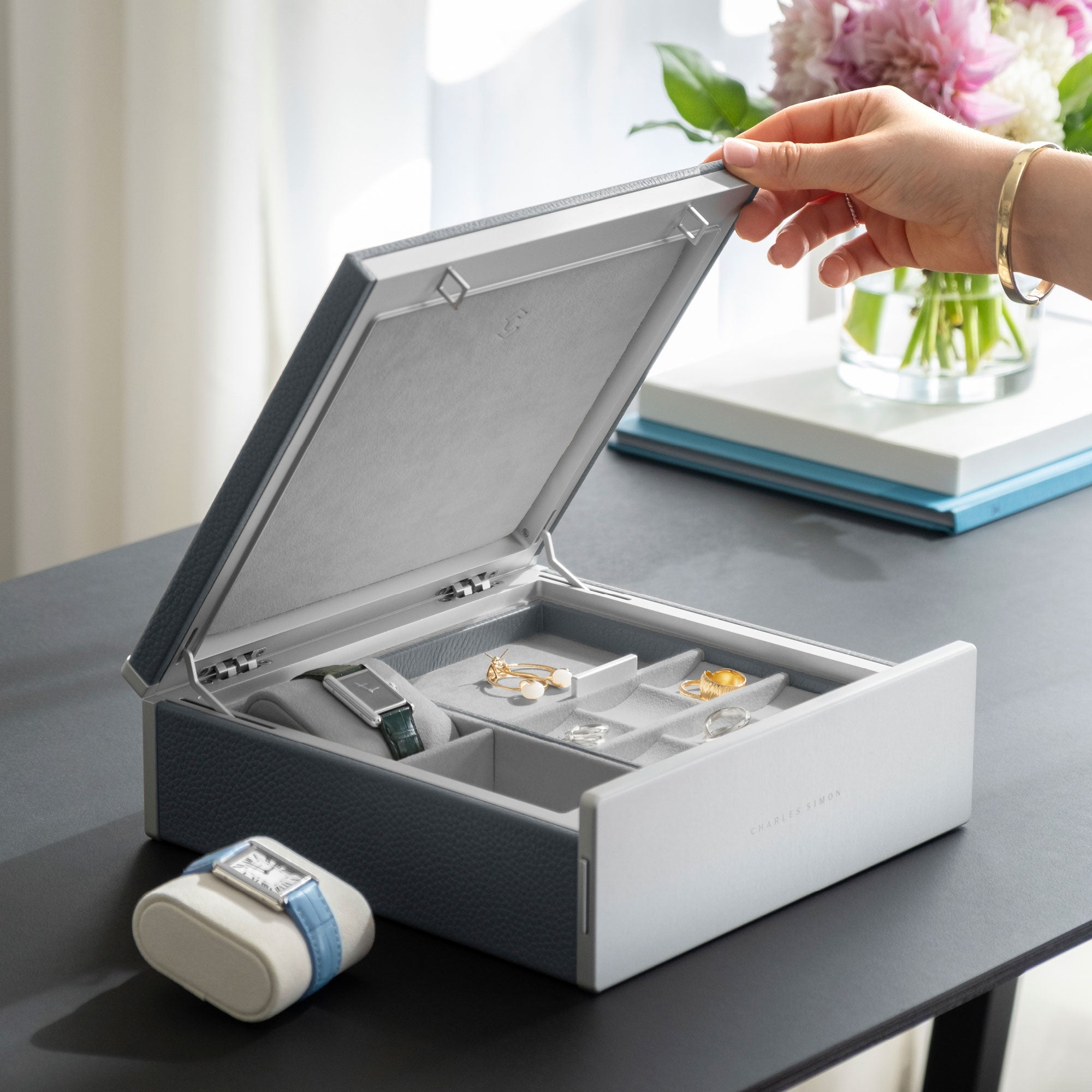 Woman opening lid of the Taylor 2 Watch and Jewelry box in cloud grey leather and fog grey interior. The inside of the Taylor 2 is holding two Cartier watches on removable watch cushions and a collection of earrings, rings, bracelets and necklaces that are organized in the jewelry boxe's mobile organization compartments.