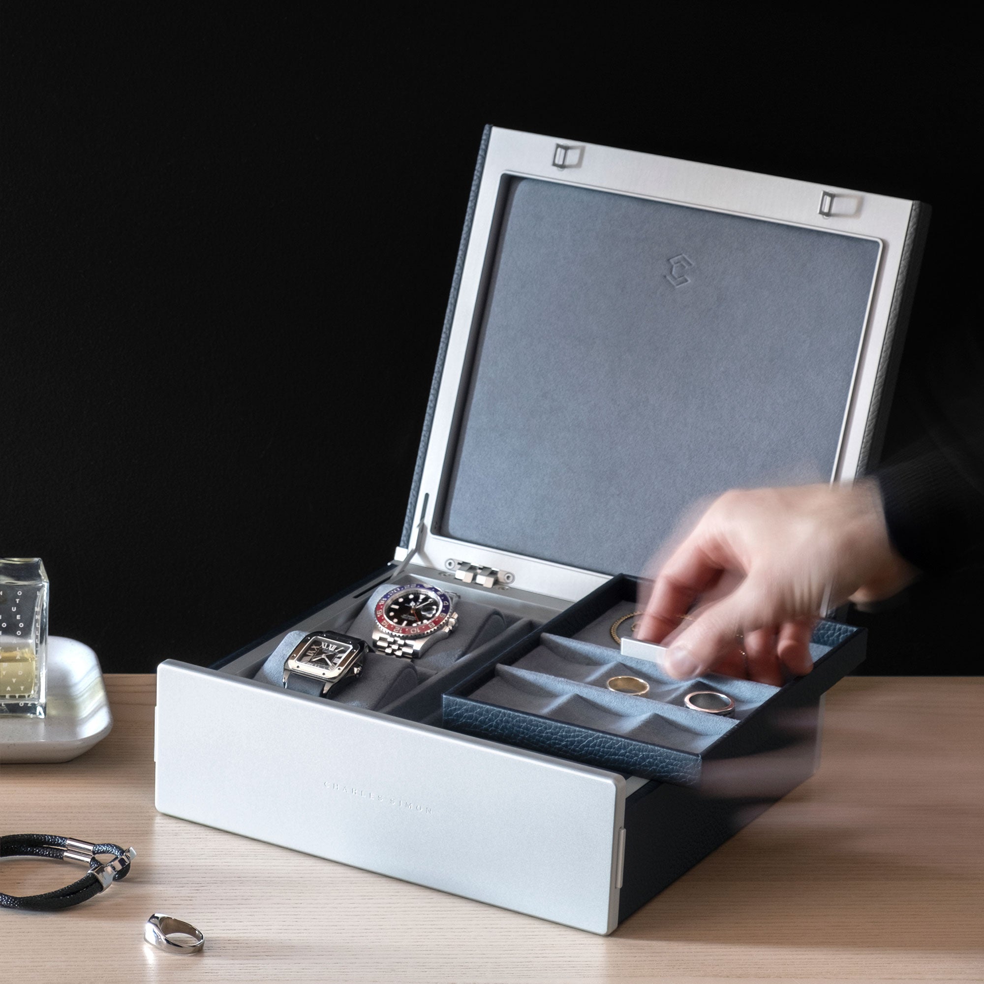 Lifestyle photo of man grabbing movable jewelry tray from the Taylor 2 Watch and Jewelry box in marine leather and smoky blue interior. The jewelry organization compartments hold gold and silver rings and two luxury watches are placed on removable watch cushions.