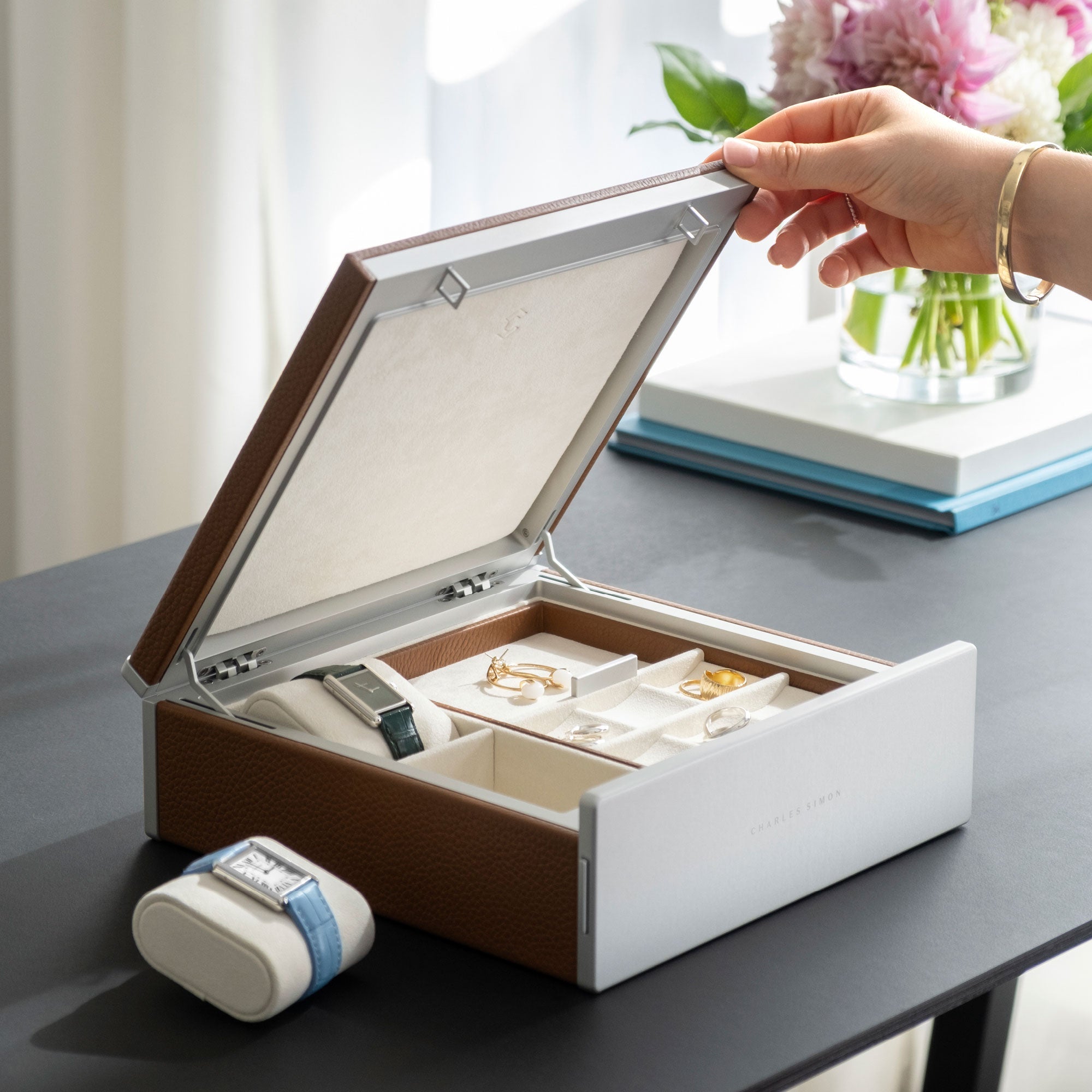 Woman closing lid of the tan leather Taylor 2 Watch and Jewelry box by Charles Simon. The watch and jewelry storage box is holding two Cartier watches and earrings, rings and a gold bracelet.