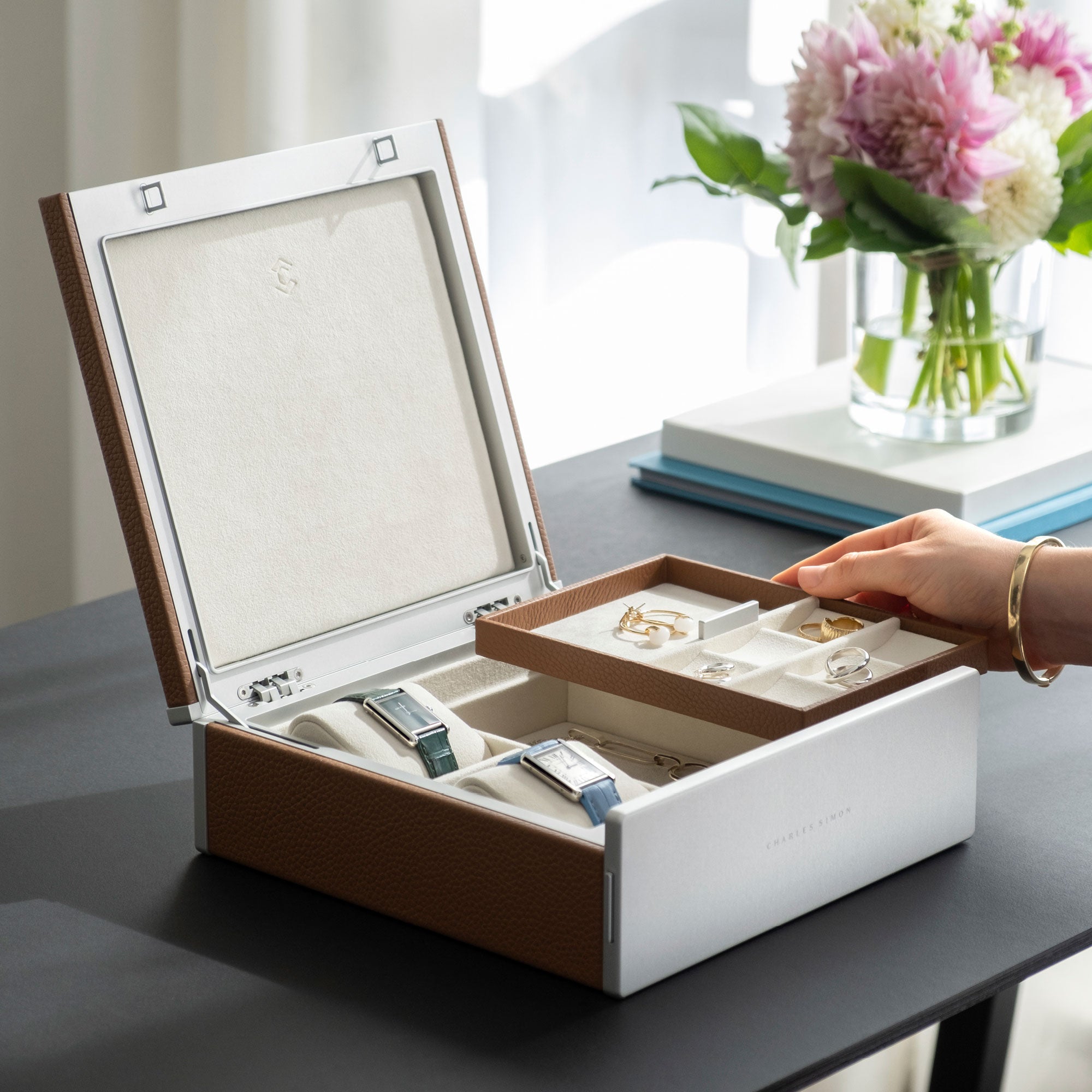 Woman grabbing jewelry tray from the Taylor 2 Watch and Jewelry box in tan leather. The jewelry storage box is displaying a fine jewelry collection of silver and gold rings and earrings, as well as a necklace.