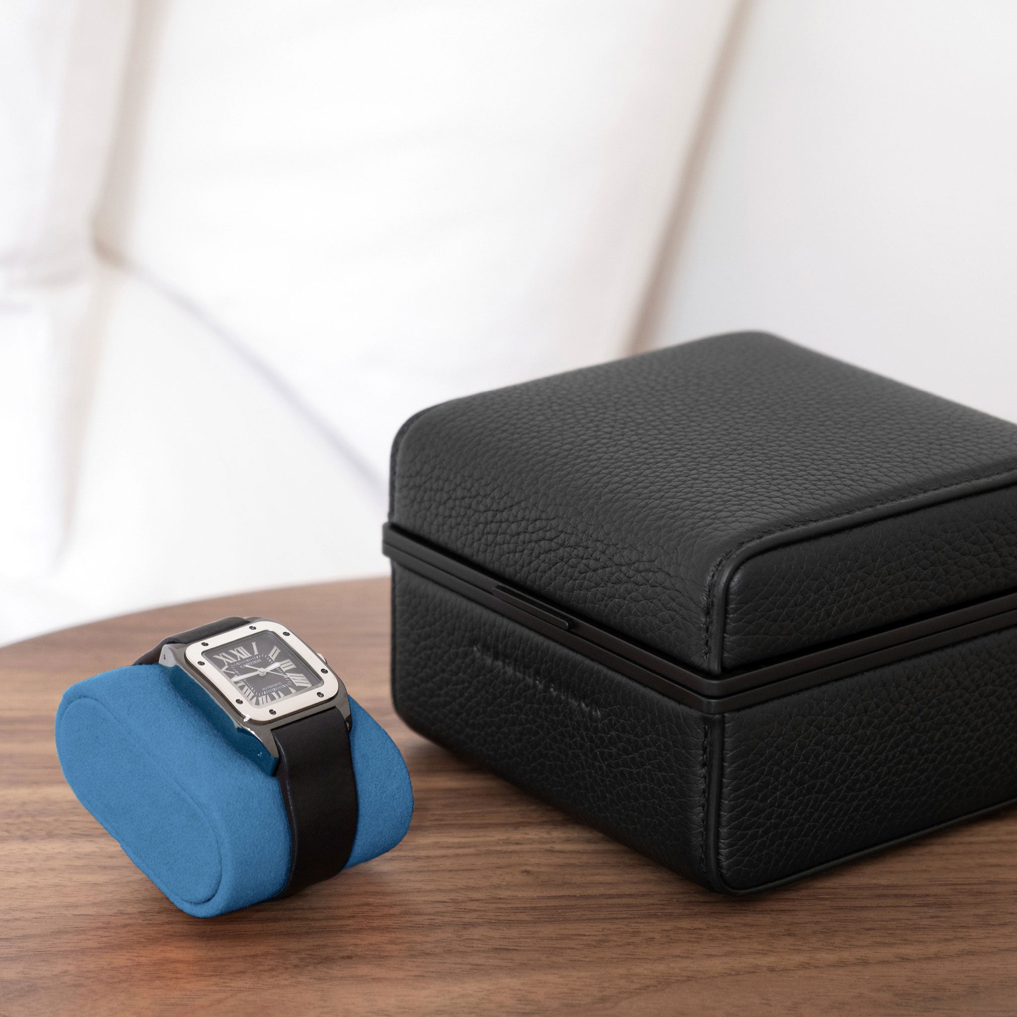 Lifestyle photo of all black Eaton 1 Watch case with contrasting cyan blue interior sitting closed on minimalist table. Cartier Santos luxury watch is placed on cyan blue removable Alcantara cushion next to closed luxury watch case
