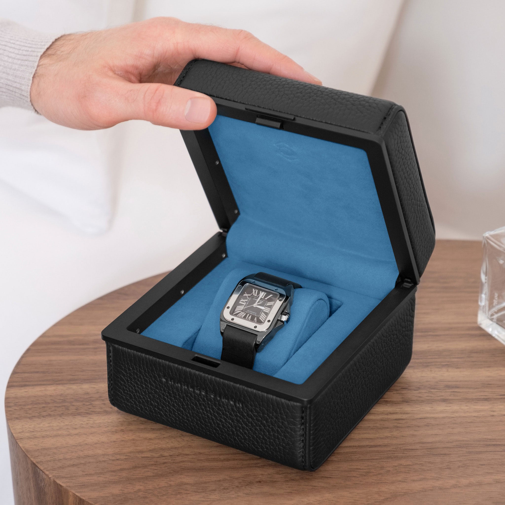 Lifestyle photo of all black Eaton 1 watch case with contrasting cyan blue interior on modern nightstand. Man is opening luxury watch case with Cartier Santos placed on cyan blue removable watch cushion.