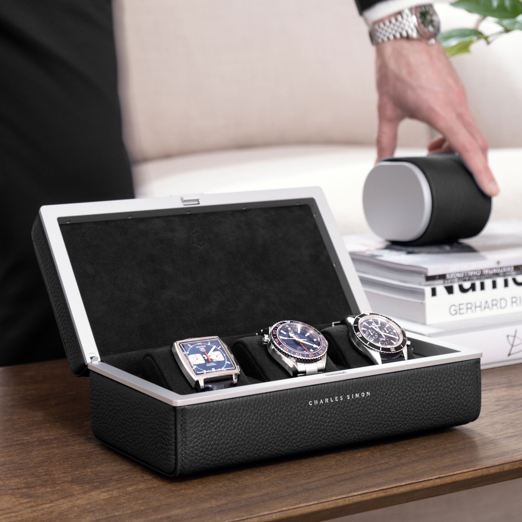 Lifestyle shot of open Eaton 3 watch case for 3 watches filled with luxury watches. Gentleman wearing Rolex is grabbing Theo watch roll placed on living room table