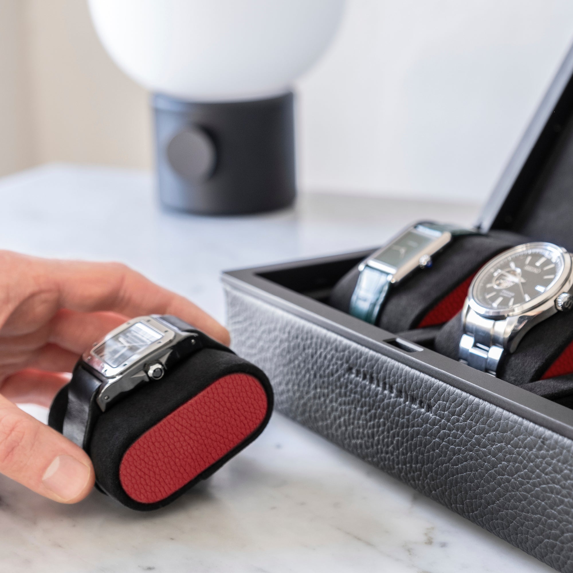 Lifestyle photo of man holding Alcantara cushion in black and contrasting red leather accents with a Cartier watch placed on it. Eaton 3 watch case in black and red leather accents is placed on the right