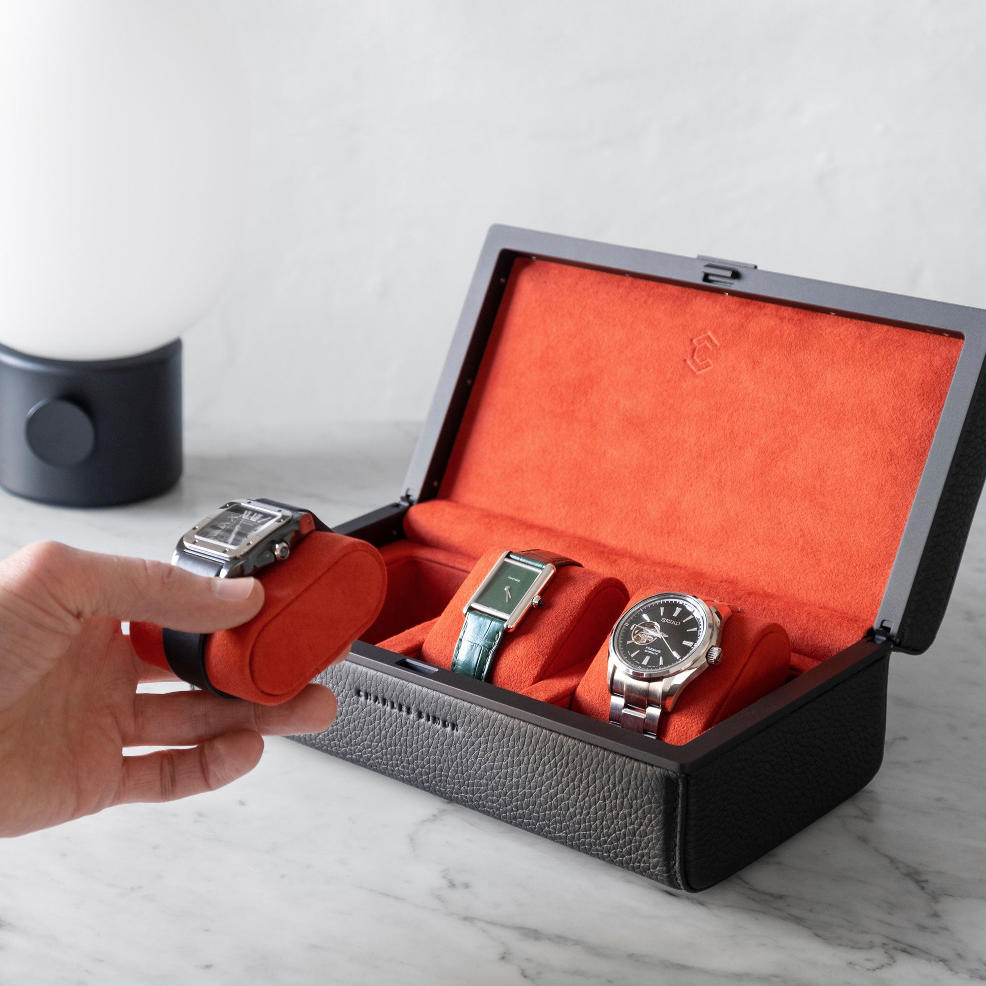 Lifestyle shot of man taking Cartier watch placed on vermilion removable Alcantara cushion from a Charles Simon Eaton 3 watch case in black leather with black anodized aluminum that is holding three men’s watches