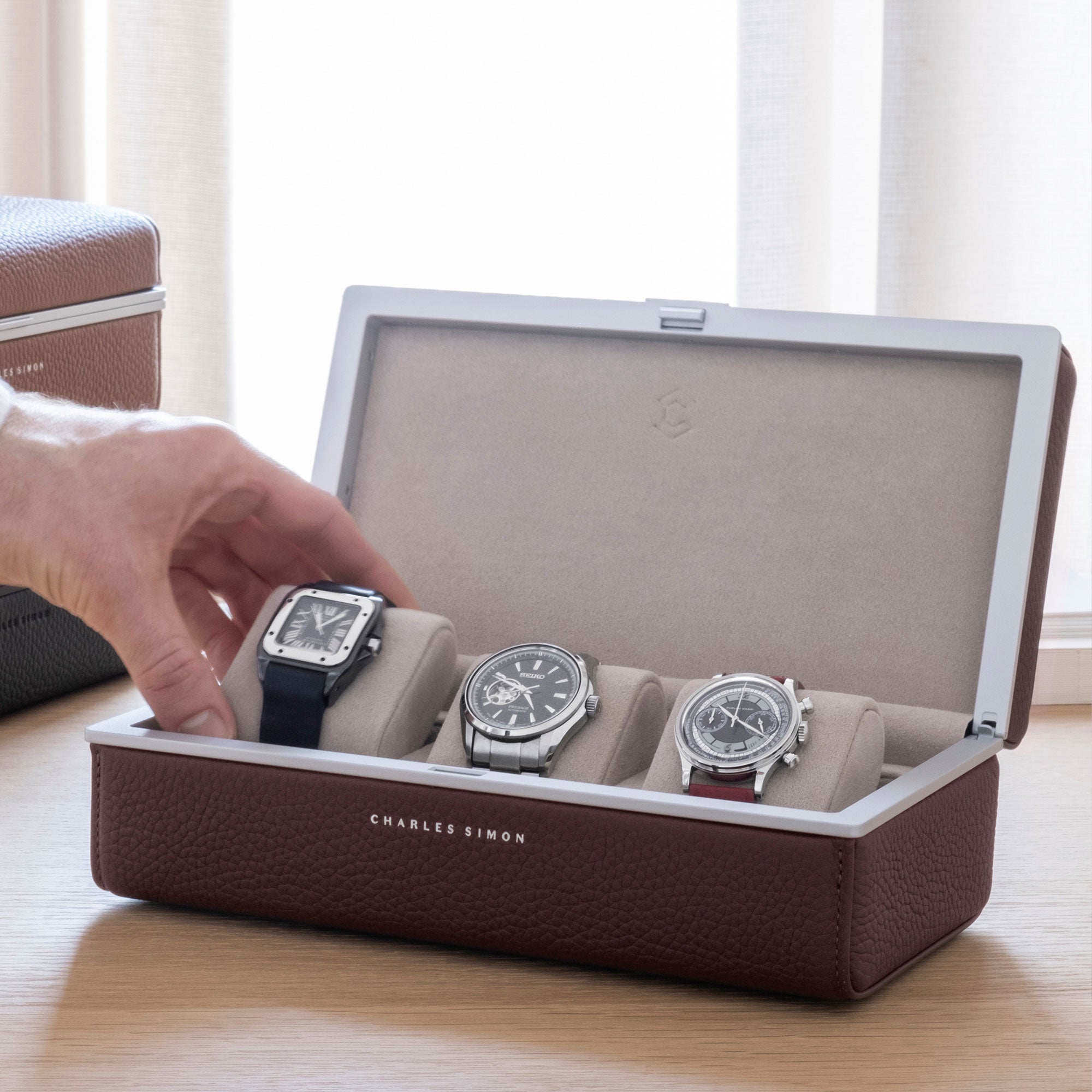 Charles Simon Eaton 3 Watch case in burgundy leather holding and displaying three luxury watches. Man is grabbing his Cartier watch showcased on a sea sand watch cushion from the handmade watch case.