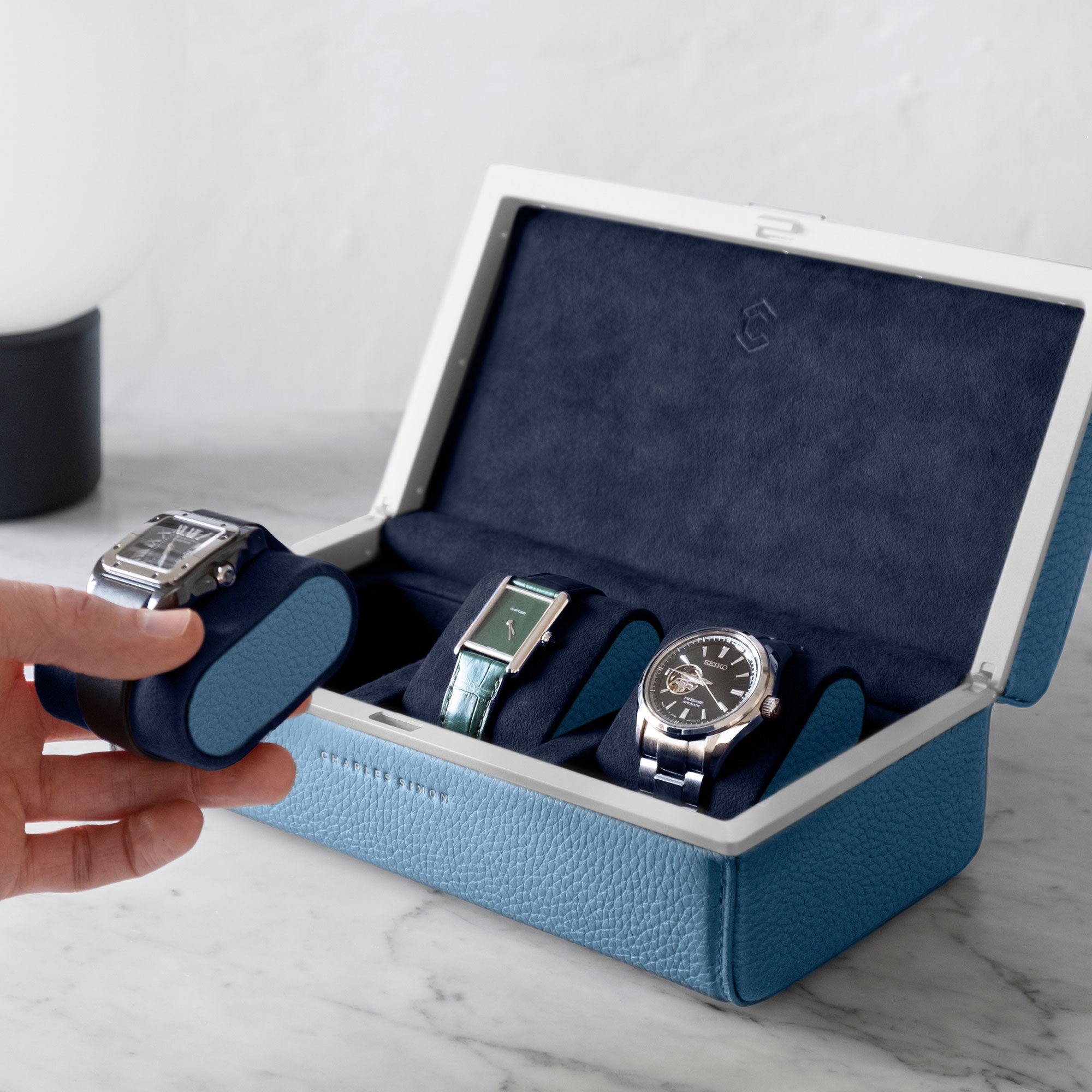 Lifestyle photo of man taking Cartier watch from the Eaton 3 watch case in sky blue leather, deep blue Alcantara interior and sky blue leather accents. The handmade watch case is holding a watch collection of 3 luxury watches while standing on a marble table