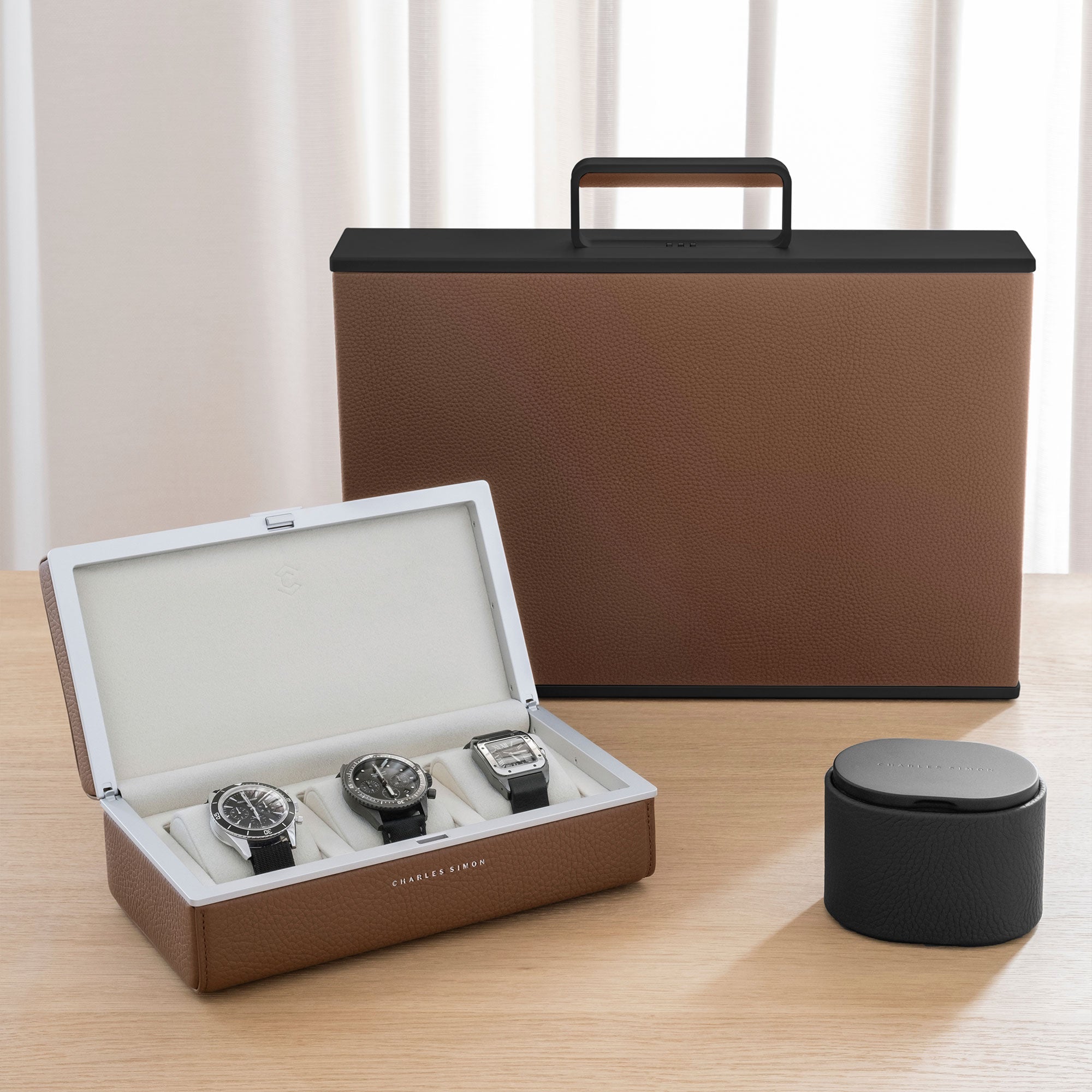 Collection of Charles Simon luxury watch accessories with open Eaton 3 watch case holding luxury watches on the left, Theo watch roll on the right and tan leather Mackenzie watch briefcase with black casing in the center of the background