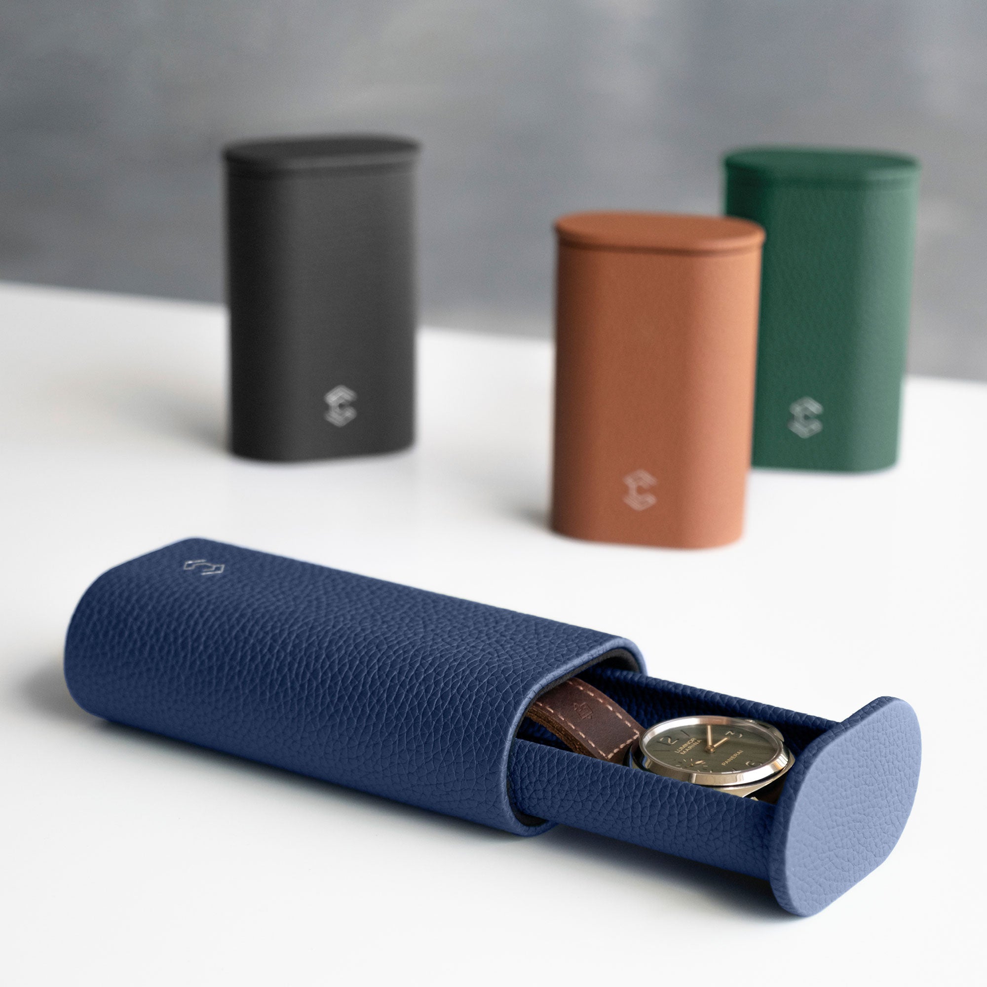 Open Oscar 1 Tall Leather Watch roll in sapphire leather holding a men's watch with a leather strap. Three closed Oscar Watch rolls in different shades of Taurillon leather are placed standing upright in the background.