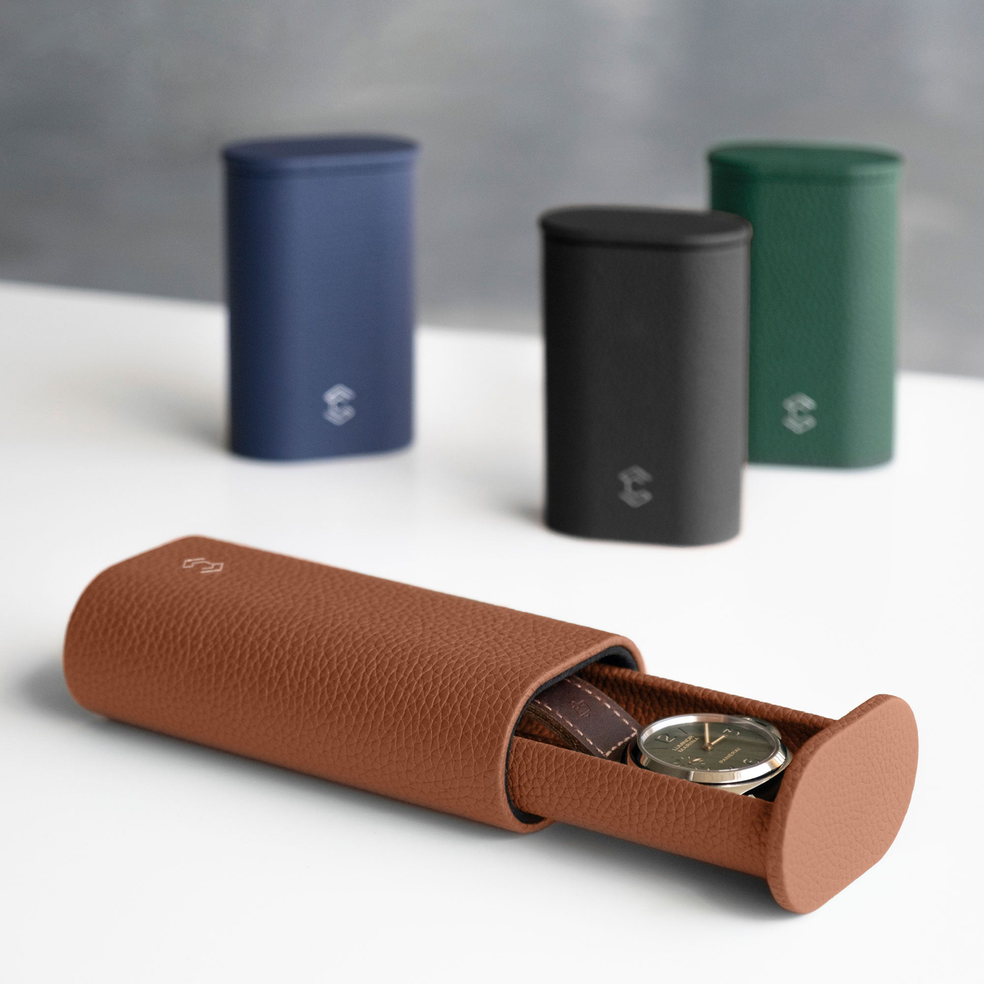 Photo of open Oscar 1 Tall Leather Watch roll in tan leather holding a luxury watch with a leather watch strap. Closed Oscar Watch rolls in different shades of French leather are placed standing upright in the background. 