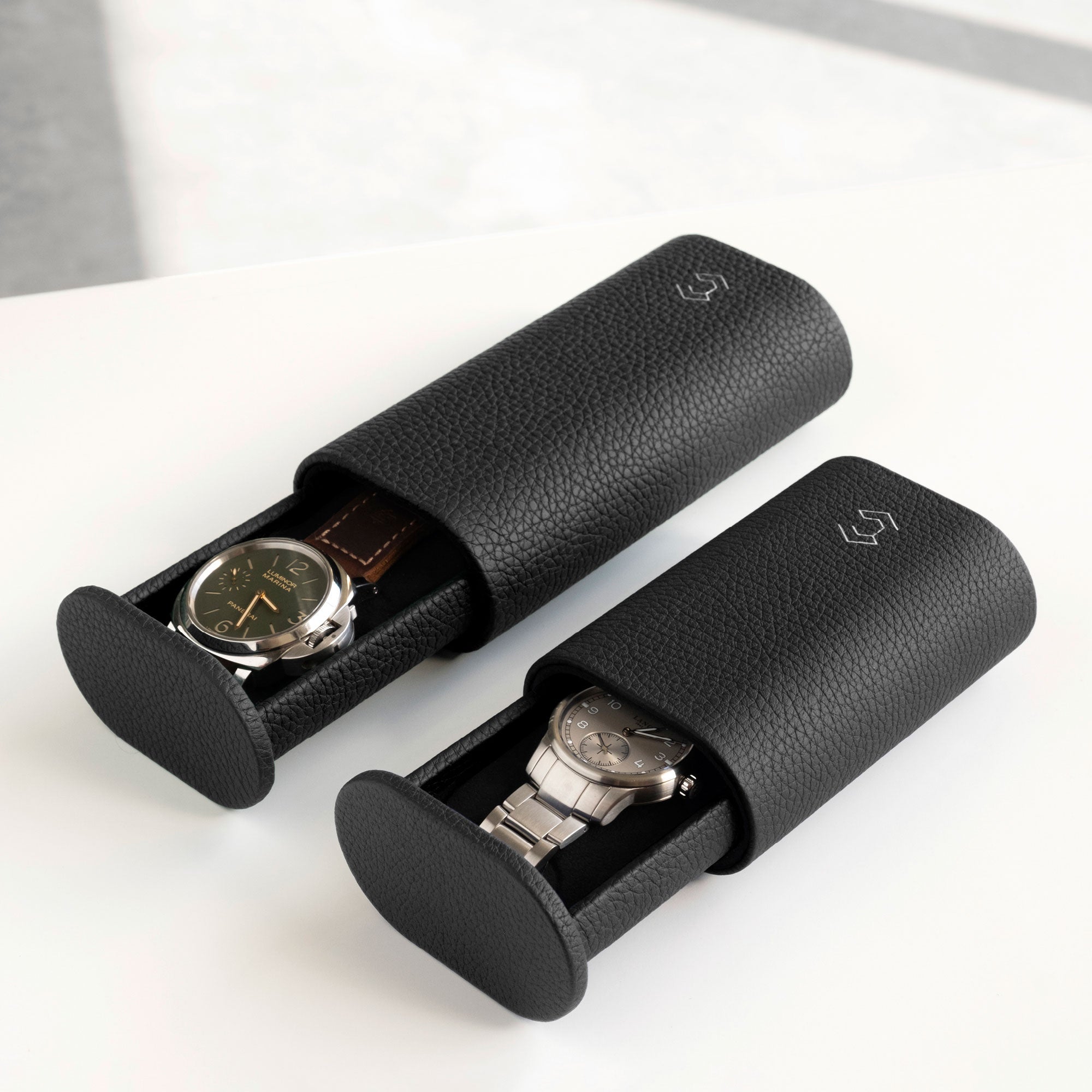 Lifestyle image of Oscar Tall watch roll in black leather on the left, holding a watch with a leather strap and a half opened Oscar Watch roll for watches with metal bracelets on the right