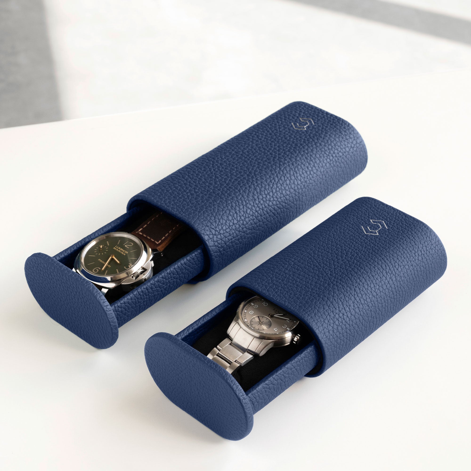 Lifestyle image of Oscar Tall watch roll in sapphire leather on the left, holding a watch with a leather strap and a half opened Oscar Watch roll in sapphire for watches with metal bracelets on the right