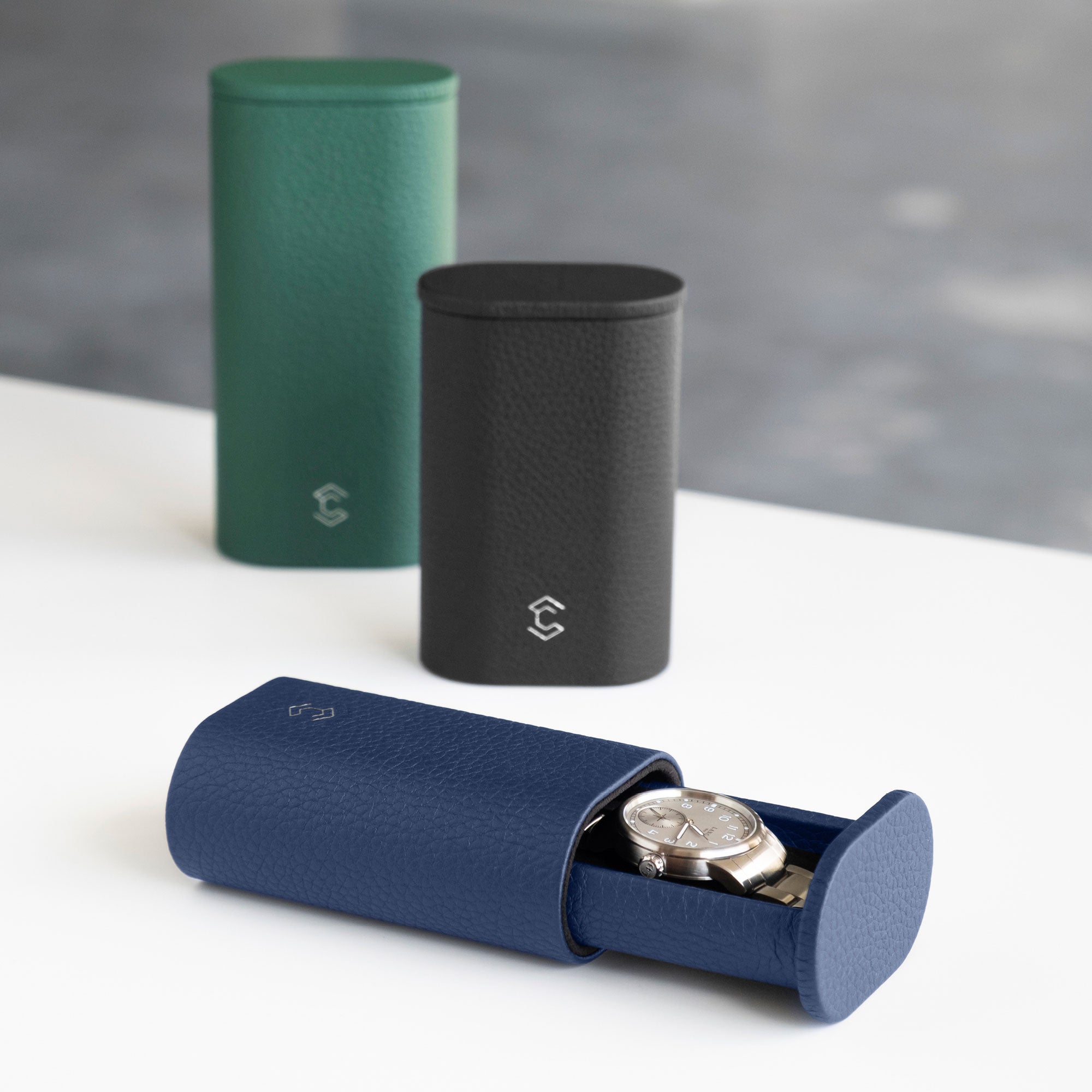 Photo of open Oscar 1 Leather Watch roll in sapphire leather holding a luxury watch with a metal bracelet. Closed Oscar Watch rolls in emerald and black French leather are placed standing upright in the background.