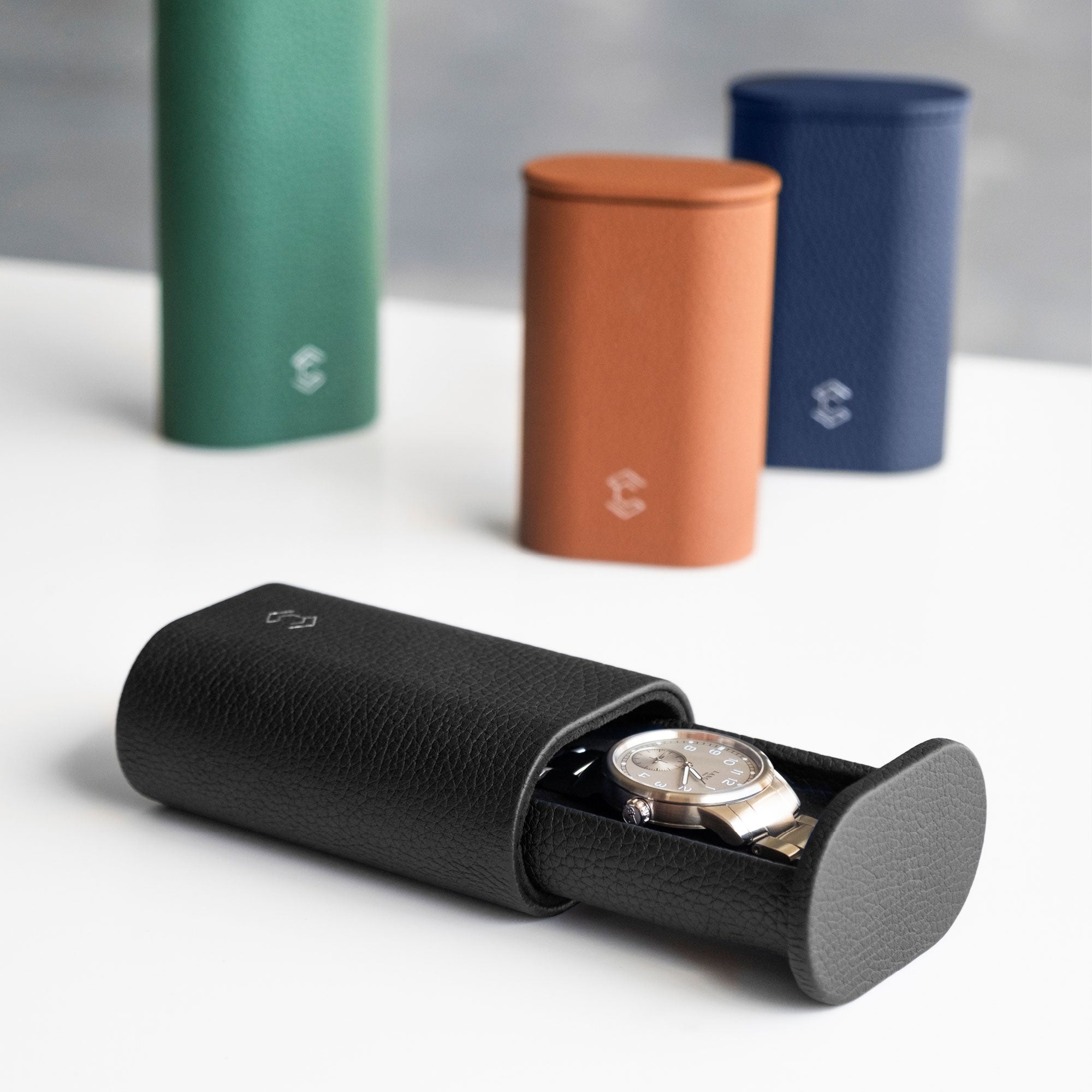 Lifestyle photo of open black Oscar Watch roll holding a luxury watch with a metal bracelet in the foreground. Three closed, standing leather watch rolls are placed in the background in tan, sapphire and emerald leather. 