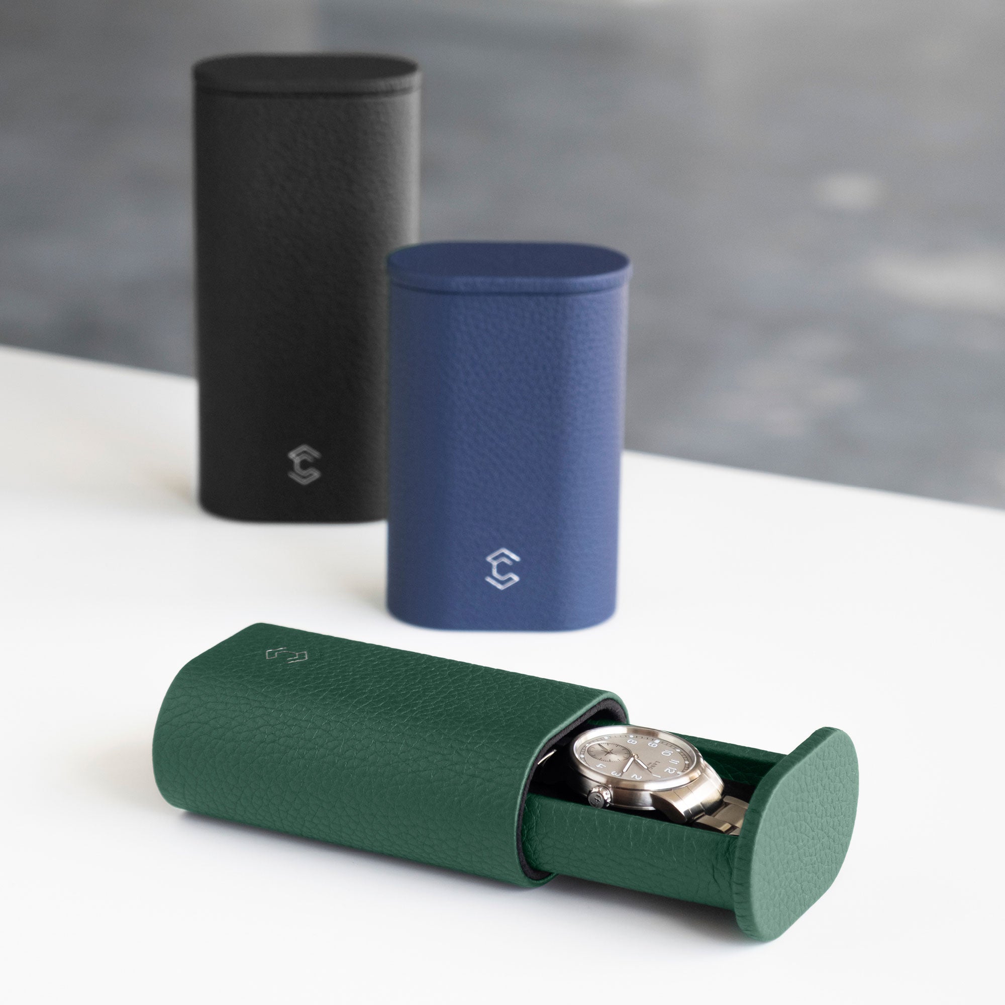 Photo of open Oscar 1 Tall Watch roll in emerald leather holding a luxury watch with a metal bracelet. Two Oscar Watch rolls in different shades of French leather are placed standing upright in the background.