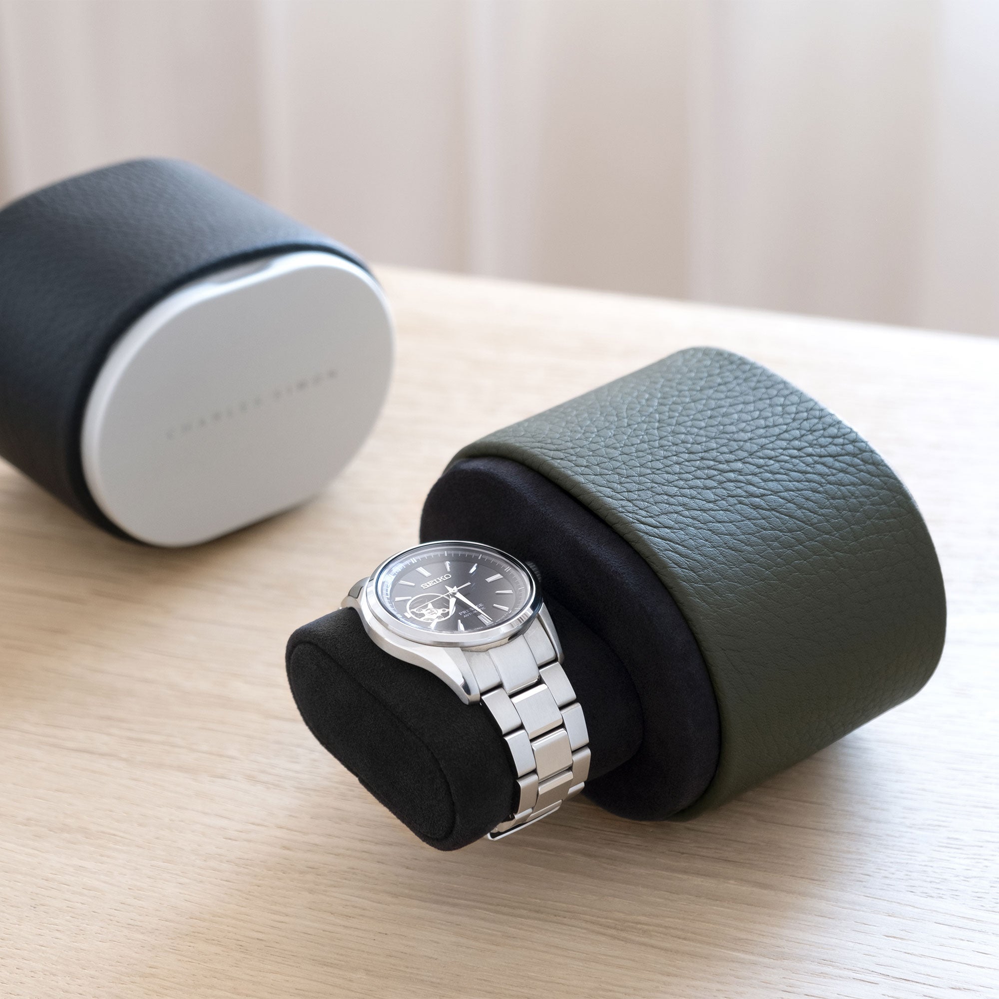 Lifestyle shot of open khaki leather Theo travel watch roll holding one watch on black Alcantara watch cushion. Closed black Theo luxury watch roll is placed in the background.