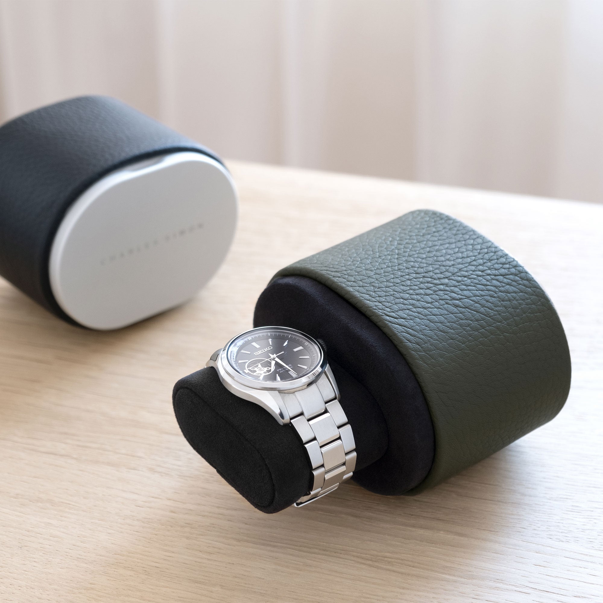 Lifestyle shot of open khaki leather Theo watch roll holding one watch on black Alcantara watch cushion. Closed black Theo luxury watch roll is placed in the background.