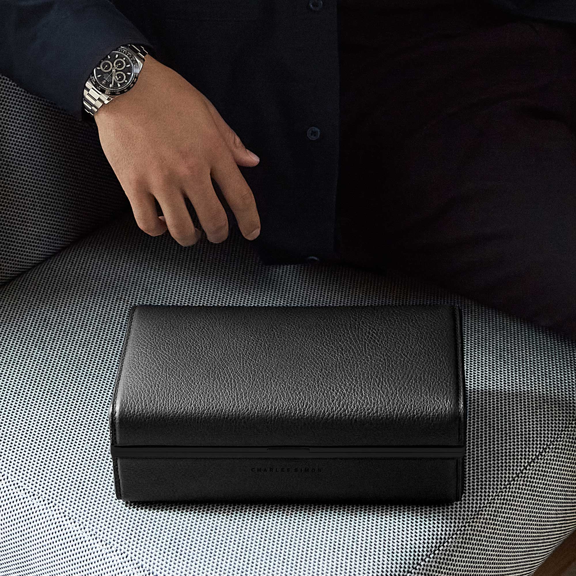 Eaton 3 watch case in all black leather, anodized aluminum and black Alcantara placed on grey couch with gentleman's hand sporting men's luxury watch hovering above