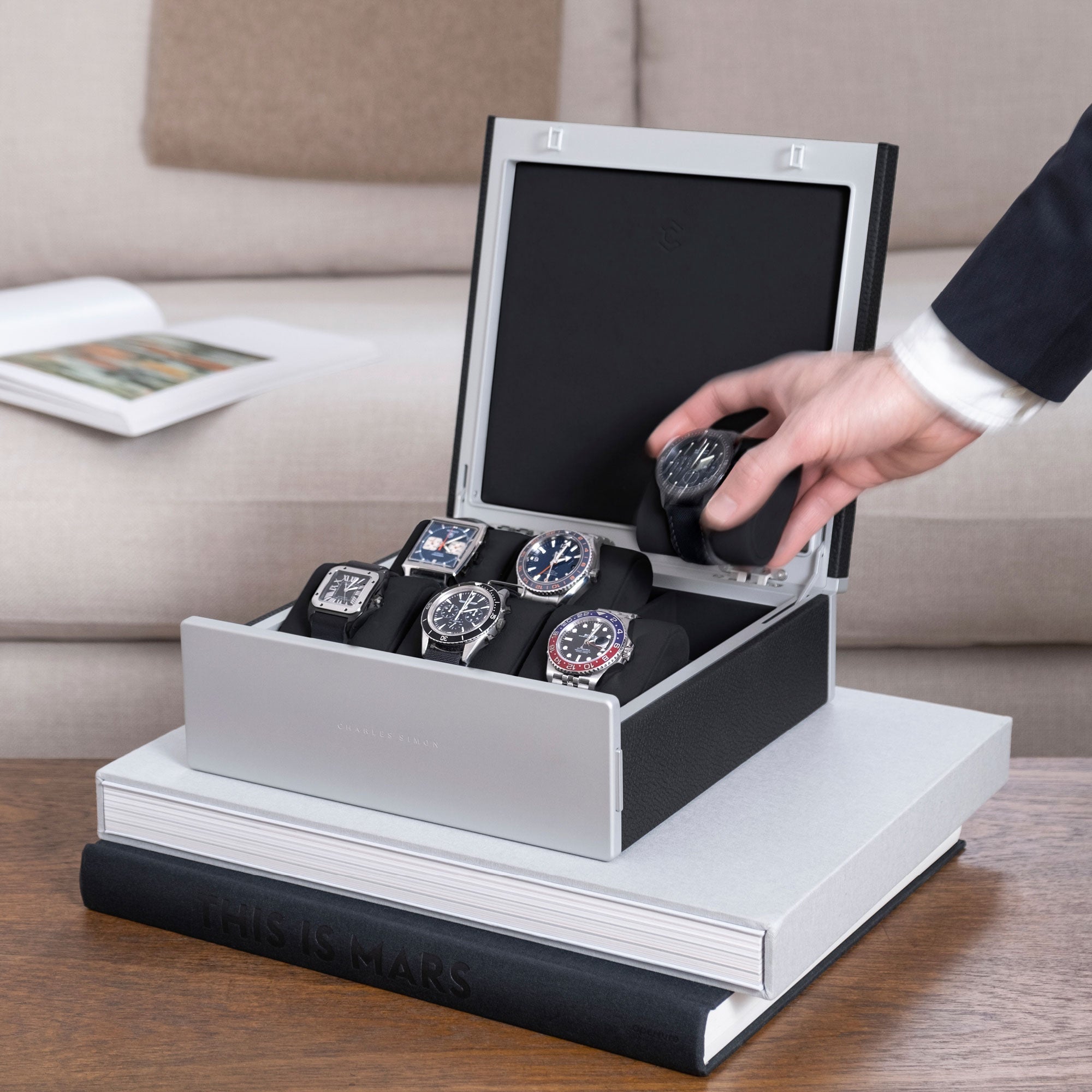 Lifestyle shot of black Spence watch box holding 6 luxury men's watches includng Rolex, Omega, Cartier, Jaeger Lecoultre. Business man is taking watch placed on black Alcantara cushion from watch box