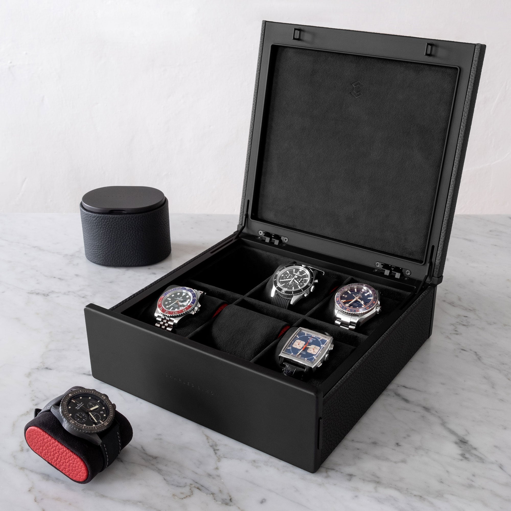 Lifestyle photo of open Spence watch box in black French leather storing luxury men's watches from Rolex, Omega, Jaeger Lecoultre and Tag Heuer. Omega Seamaster is placed in front of designer watch box on black watch cushion with red leather sides. Closed Theo watch roll in all black is placed in the background.