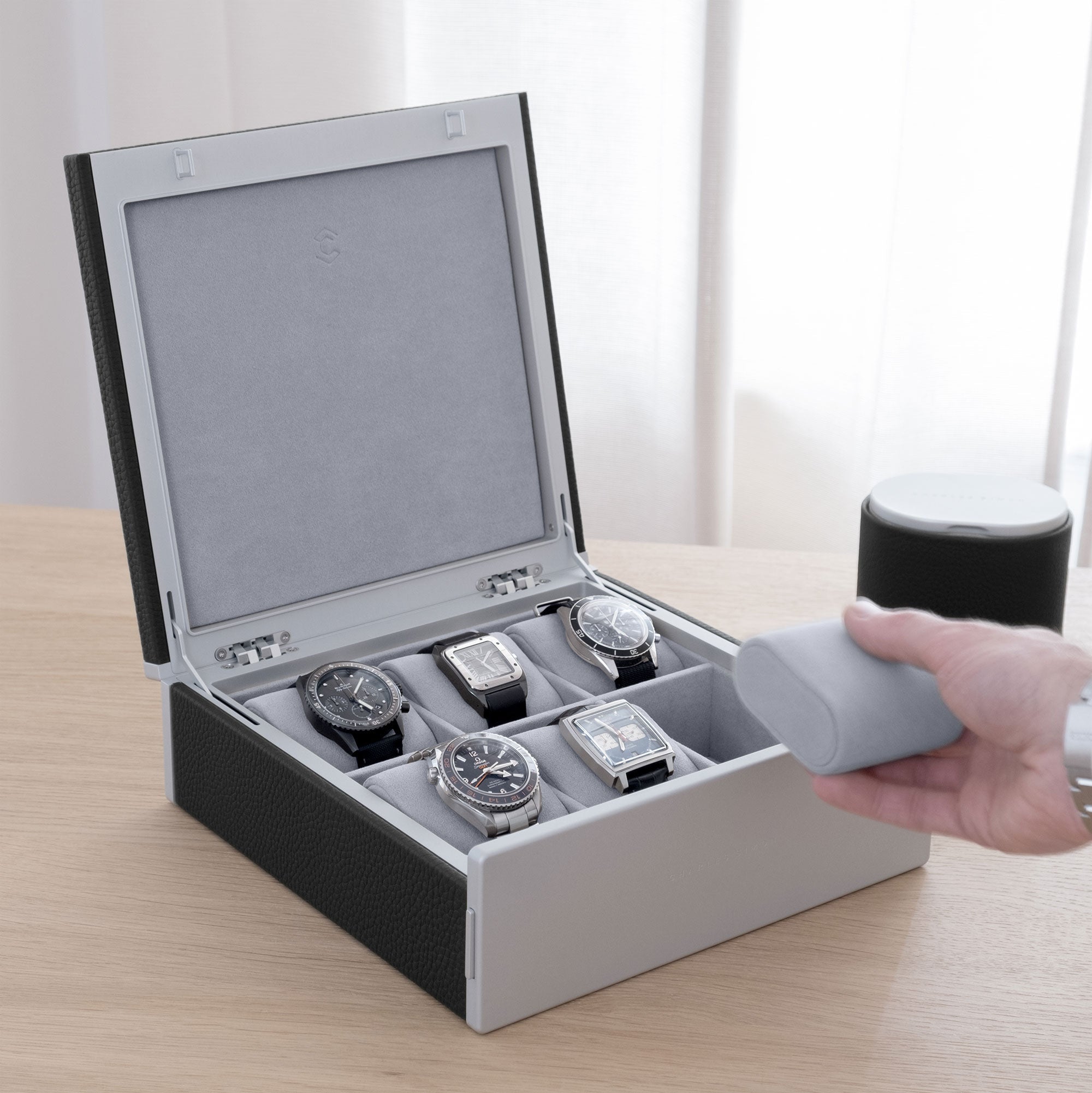 Luxury lifestyle shot of Spence watch box made from black leather for 6 watches filled with men's luxury watches including Rolex, IWC, Jaeger Lecoultre, Cartier and Omega. Gentleman is taking removable fog grey Alcantara cushion from the Spence watch box.