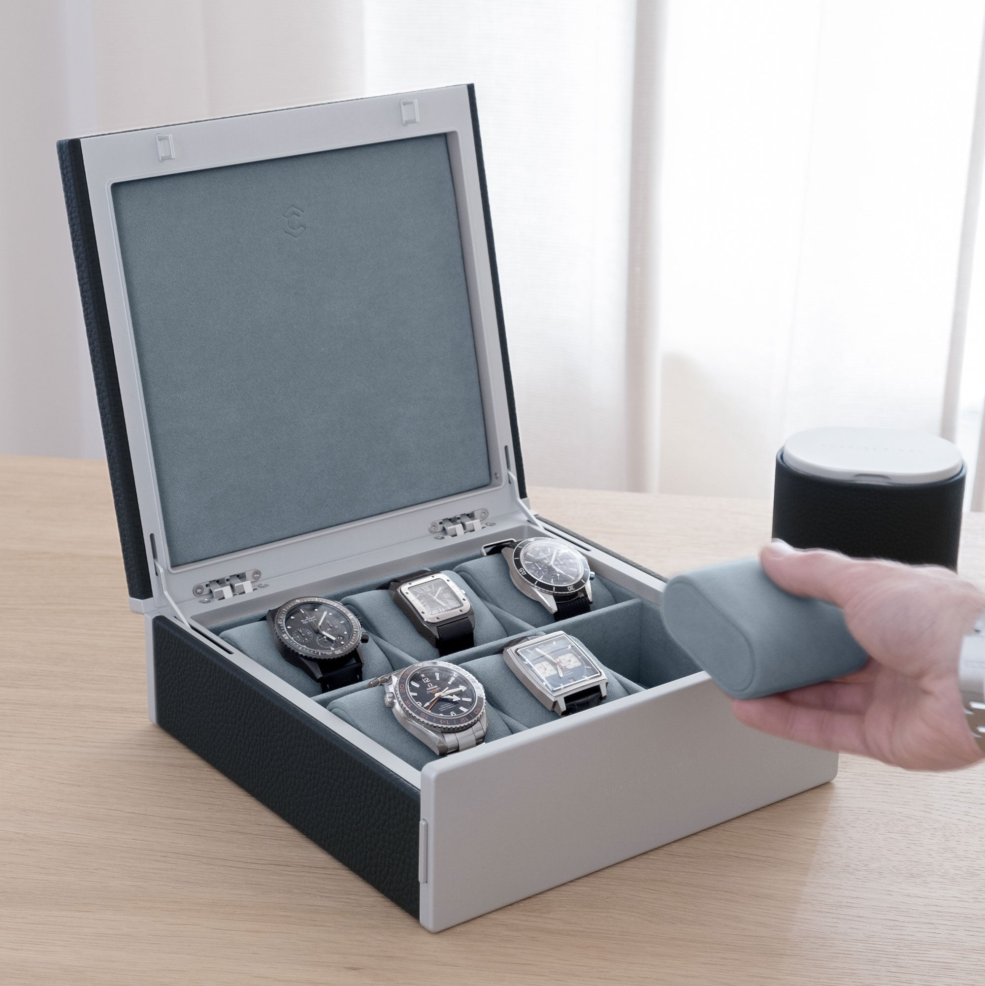 Luxury lifestyle shot of marine leather Spence watch box for 6 watches filled with men's luxury watches including Rolex, IWC, Jaeger Lecoultre, Cartier and Omega. Gentleman is taking removable Alcantara cushion from the smoky blue interior of the Spence watch box.
