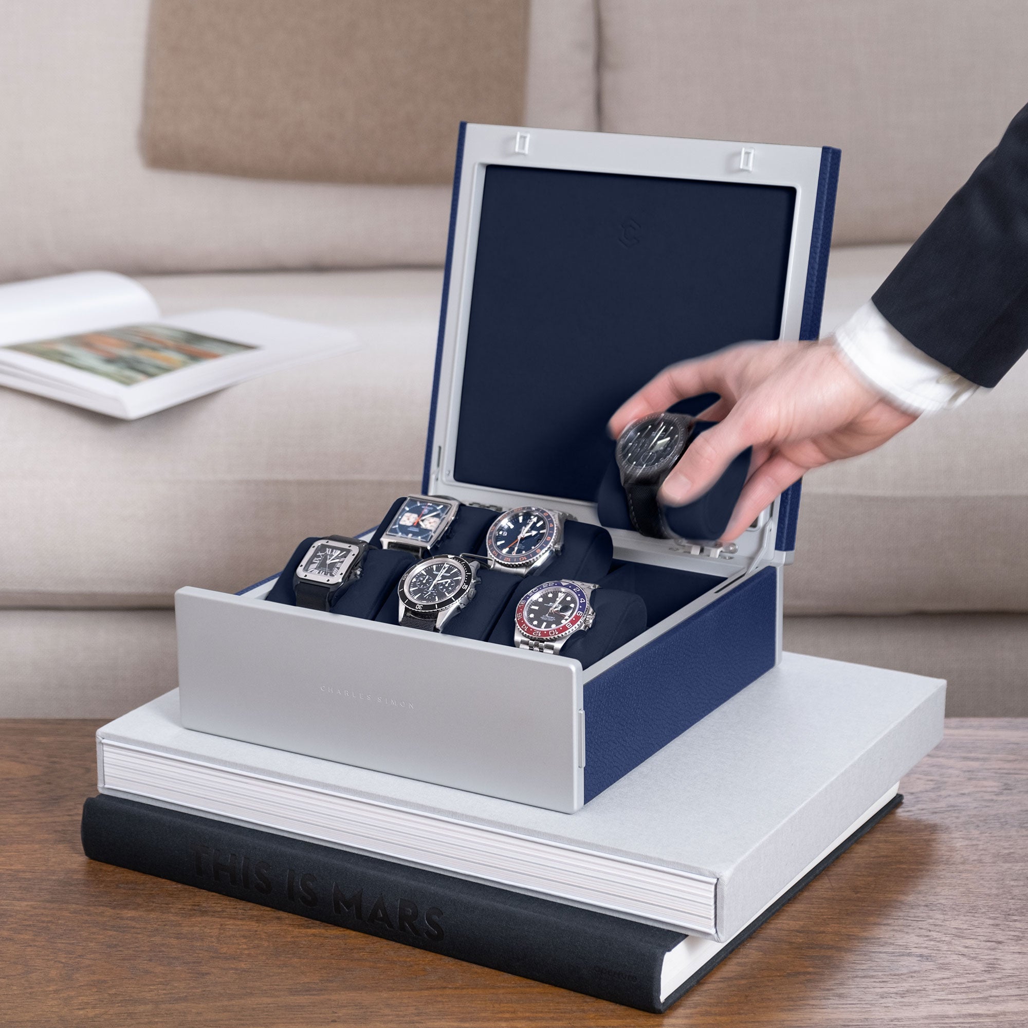 Lifestyle shot of sapphire Spence watch box holding 6 luxury men's watches includng Rolex, Omega, Cartier, Jaeger Lecoultre. Business man is taking watch placed on deep blue Alcantara cushion from watch box 