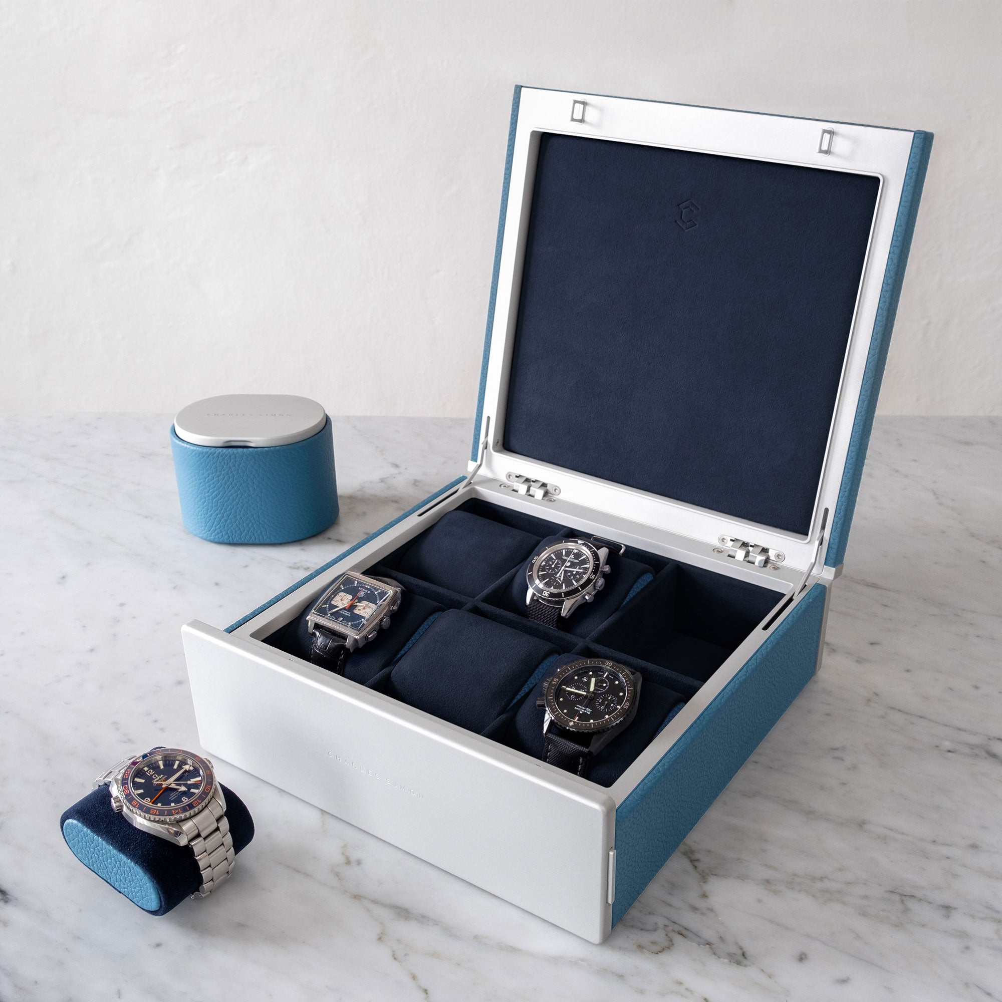 Lifestyle photo of open Spence watch box in sky blue French leather storing luxury men's watches from Rolex, Omega, Jaeger Lecoultre and Tag Heuer. Omega Seamaster is placed in front of designer watch box on watch cushion with sky blue leather sides. Closed Theo watch roll in sky blue is placed in the background.