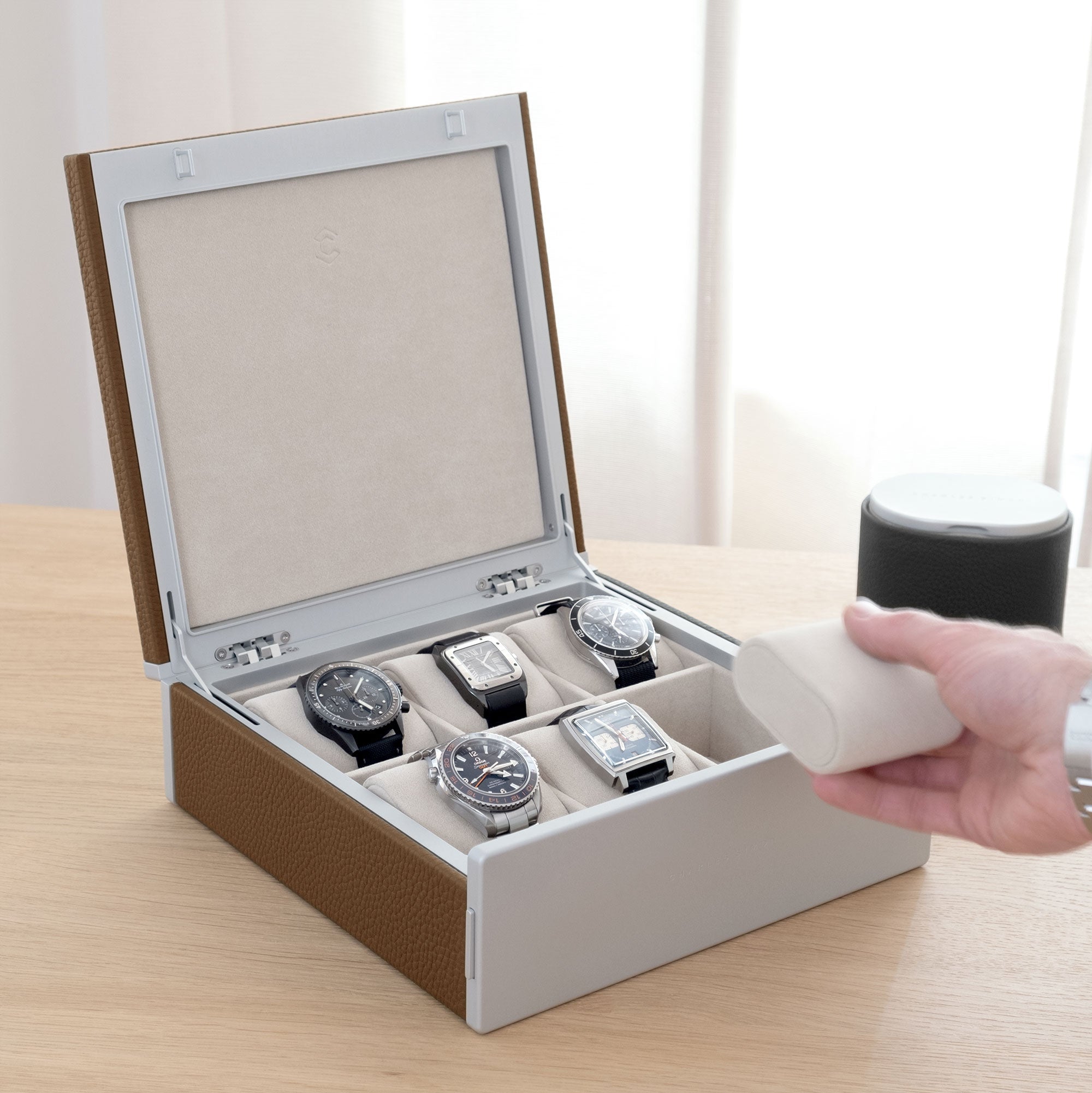 Luxury watch box for 6 watches made from tan leather, carbon fiber and anodized aluminum casing and Alcantara interior. Gentleman is grabbing removable Alcantara from handmade watch box placed in elegant environment. Watch box is filled with 5 men's luxury watches including Cartier, Rolex, Omega, Jaeger Lecoultre. 