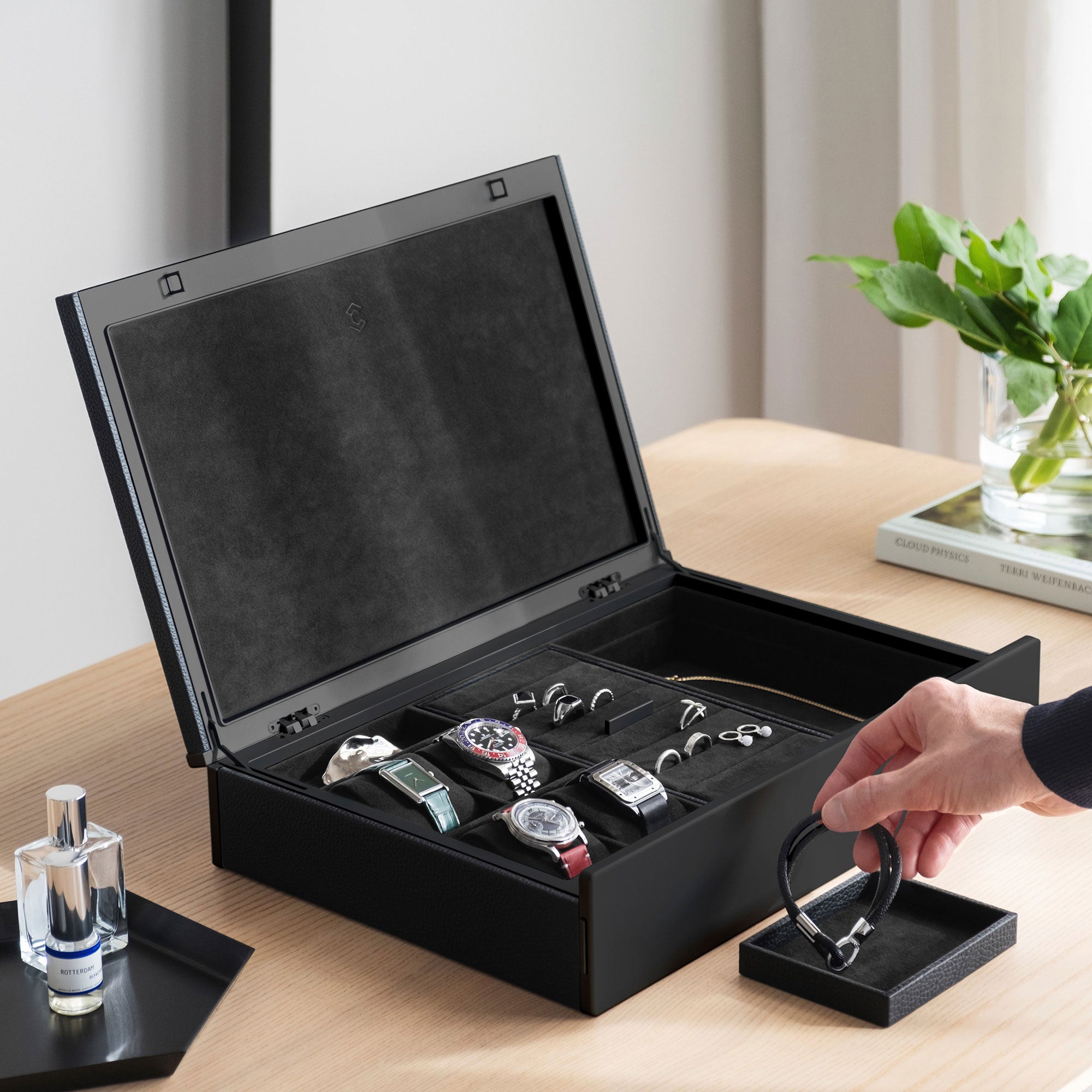 Lifestyle photo of man taking bracelet from removable jewelry tray from the all black Taylor 4 Watch and Jewelry box. The box is holding 4 watches and fine jewelry, including silver and gold rings and a necklace in a modern apartment setting. 