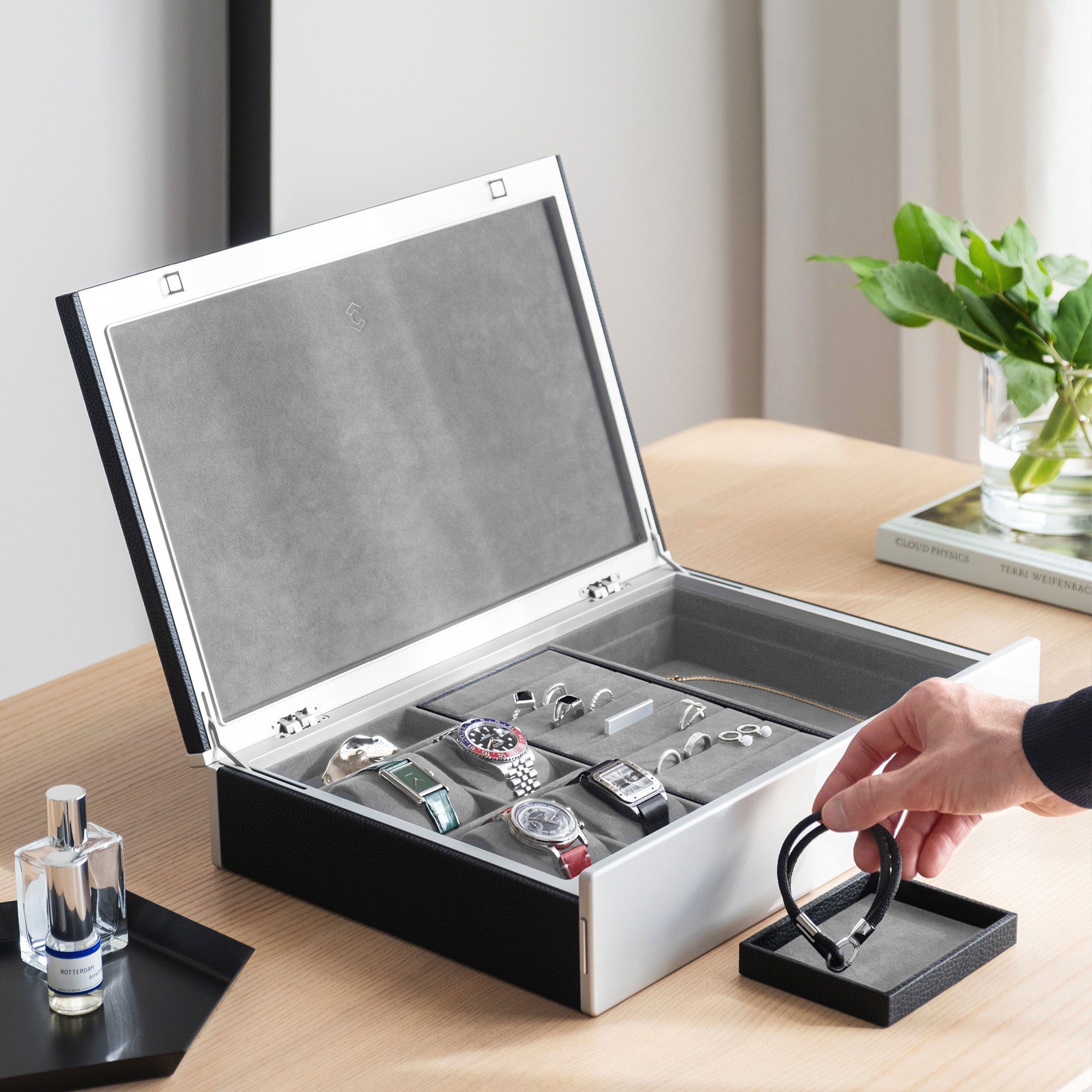 Lifestyle photo of man taking bracelet from removable jewelry tray from the black leather and fog grey interior Taylor 4 Watch and Jewelry box. The box is holding 4 watches and fine jewelry, including silver and gold rings and a necklace in a modern apartment setting.