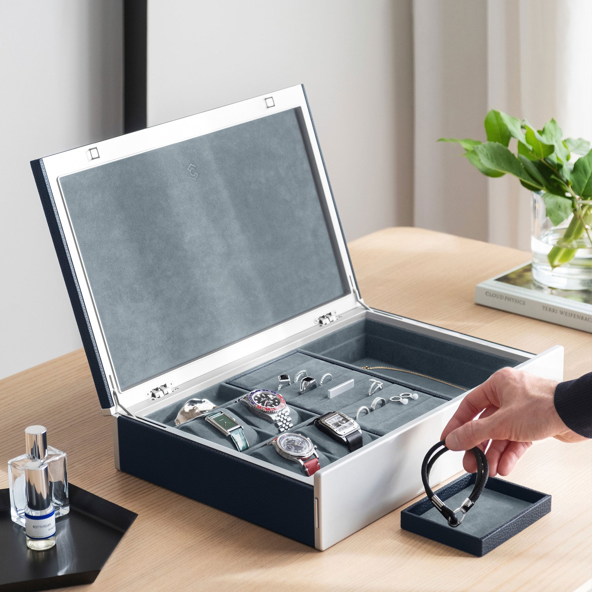 Lifestyle photo of man taking bracelet from removable jewelry tray from the marine leather and smoky blue interior Taylor 4 Watch and Jewelry box. The box is holding 4 watches and fine jewelry, including silver and gold rings and a necklace in a modern apartment setting.