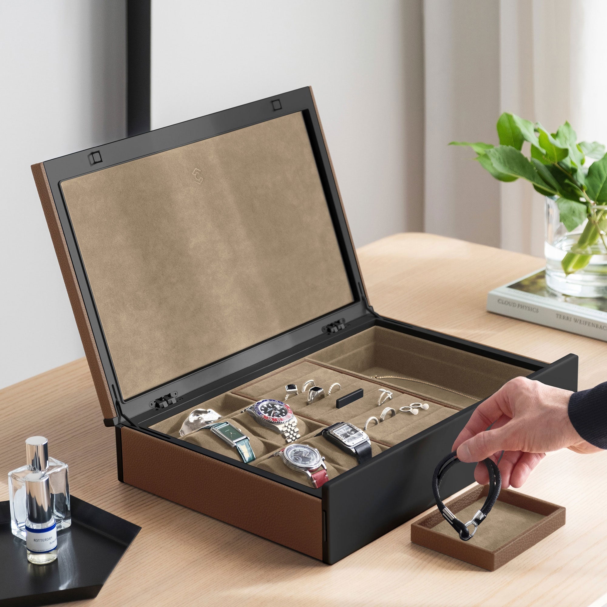 Lifestyle photo of man grabbing bracelet from the jewelry tray of the Taylor 4 Watch and Jewelry box in tan leather, black carbon fiber and anodized aluminum casing and camel interior. The jewelry organization and watch storage box is holding 4 luxury watches and a collection of rings, earrings and necklaces.