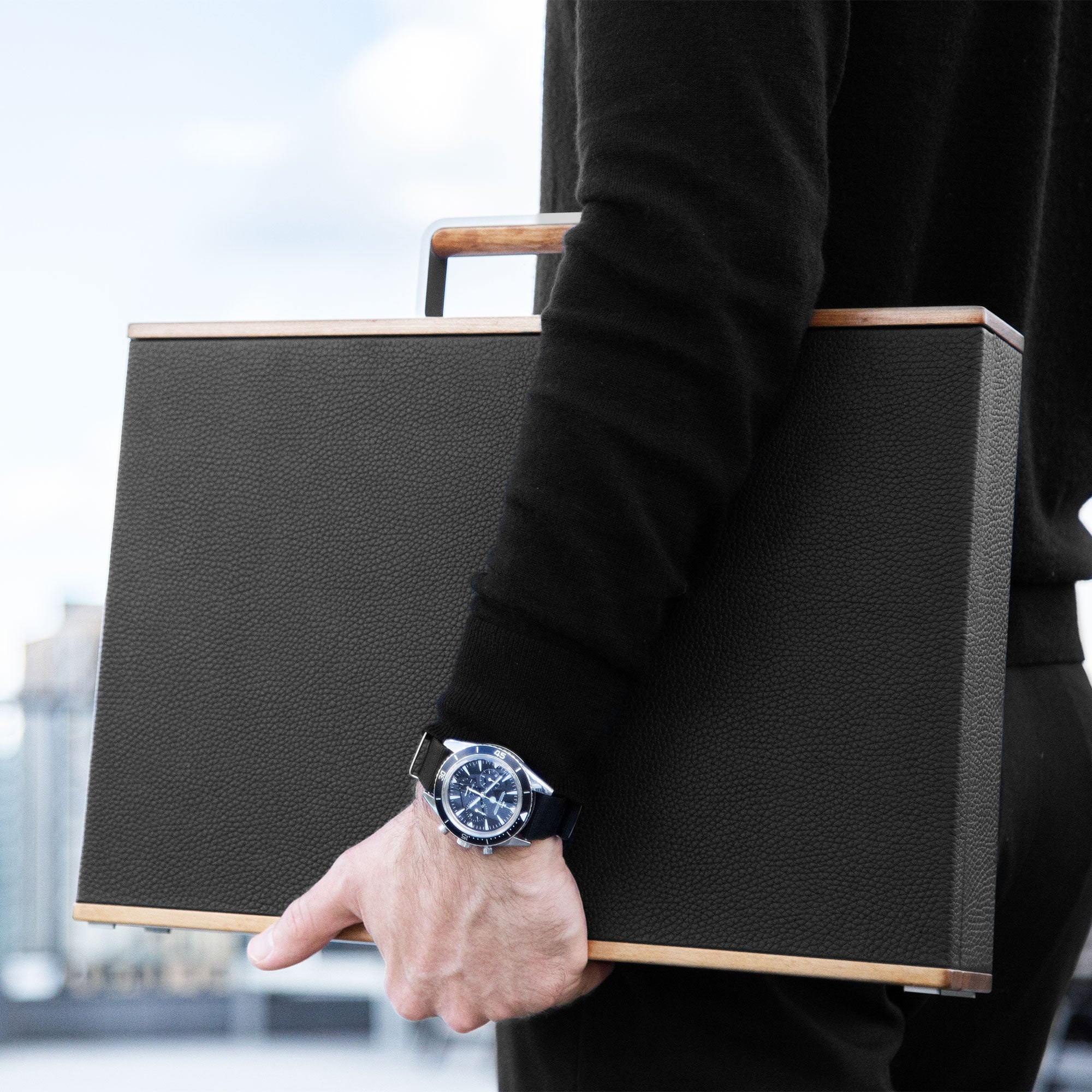 Lifestyle shot of man wearing luxury watch holding Mackenzie Original watch briefcase. Handmade from recycled wood, French leather and carbon fiber and anodized aluminum to display your watch collection